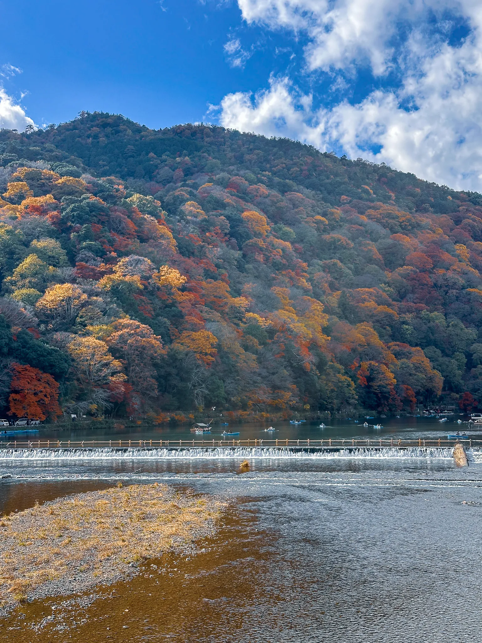 A hillside across the river turned every shade of red, orange and green, small boats on the water above a low weir and a bank of cloud overhead