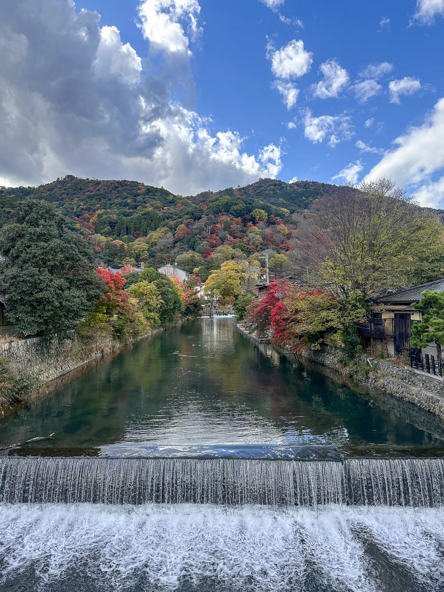 Water pouring evenly over a low weir into white foam, a green channel running back between stone banks to autumn trees, wooden houses and hills beyond