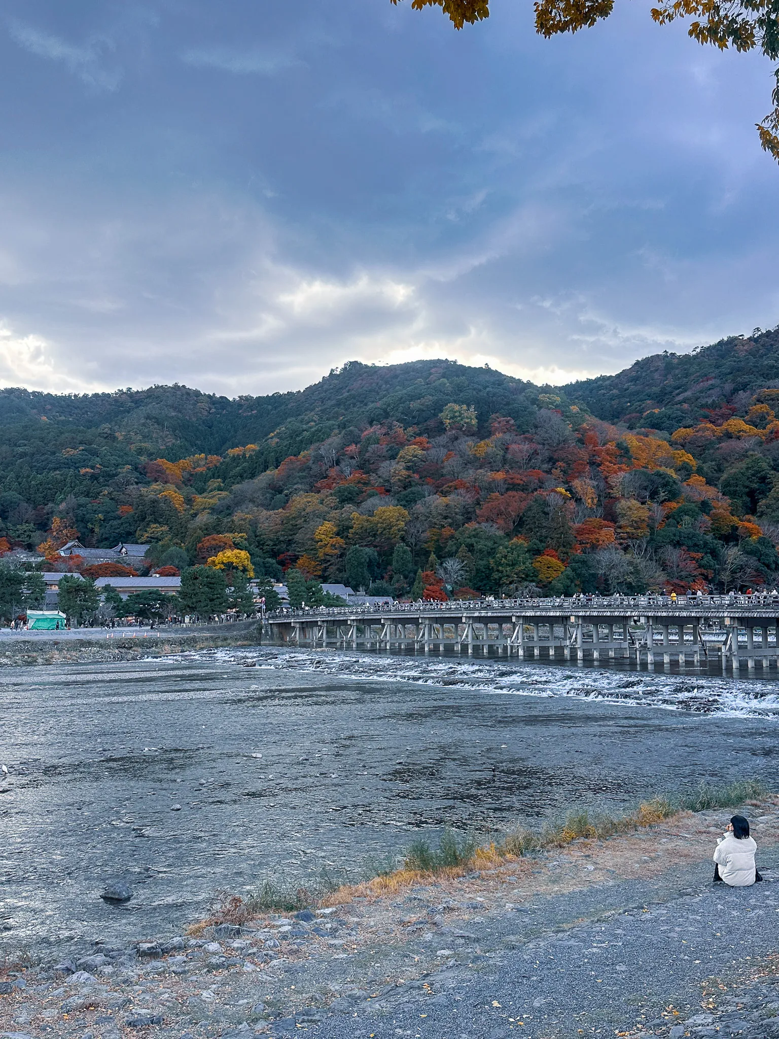 The Togetsukyo bridge crowded end to end under an autumn hillside at dusk, one person in a white coat sitting alone on the gravel bank in the foreground