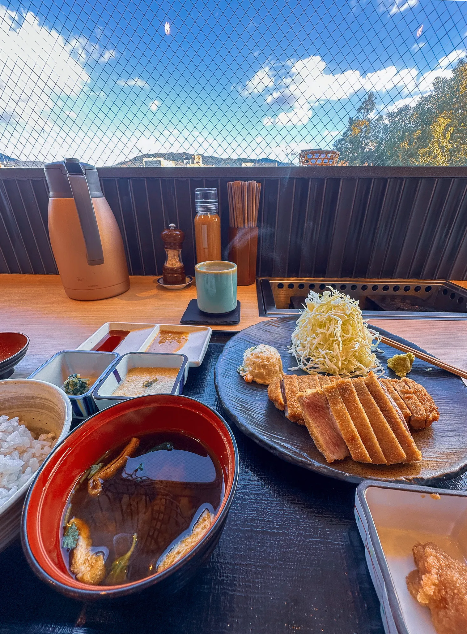 A beef katsu set on a counter facing out to the hills - sliced breaded cutlet still pink inside, a heap of shredded cabbage, a small grill to finish it on, miso soup, rice and dipping sauces