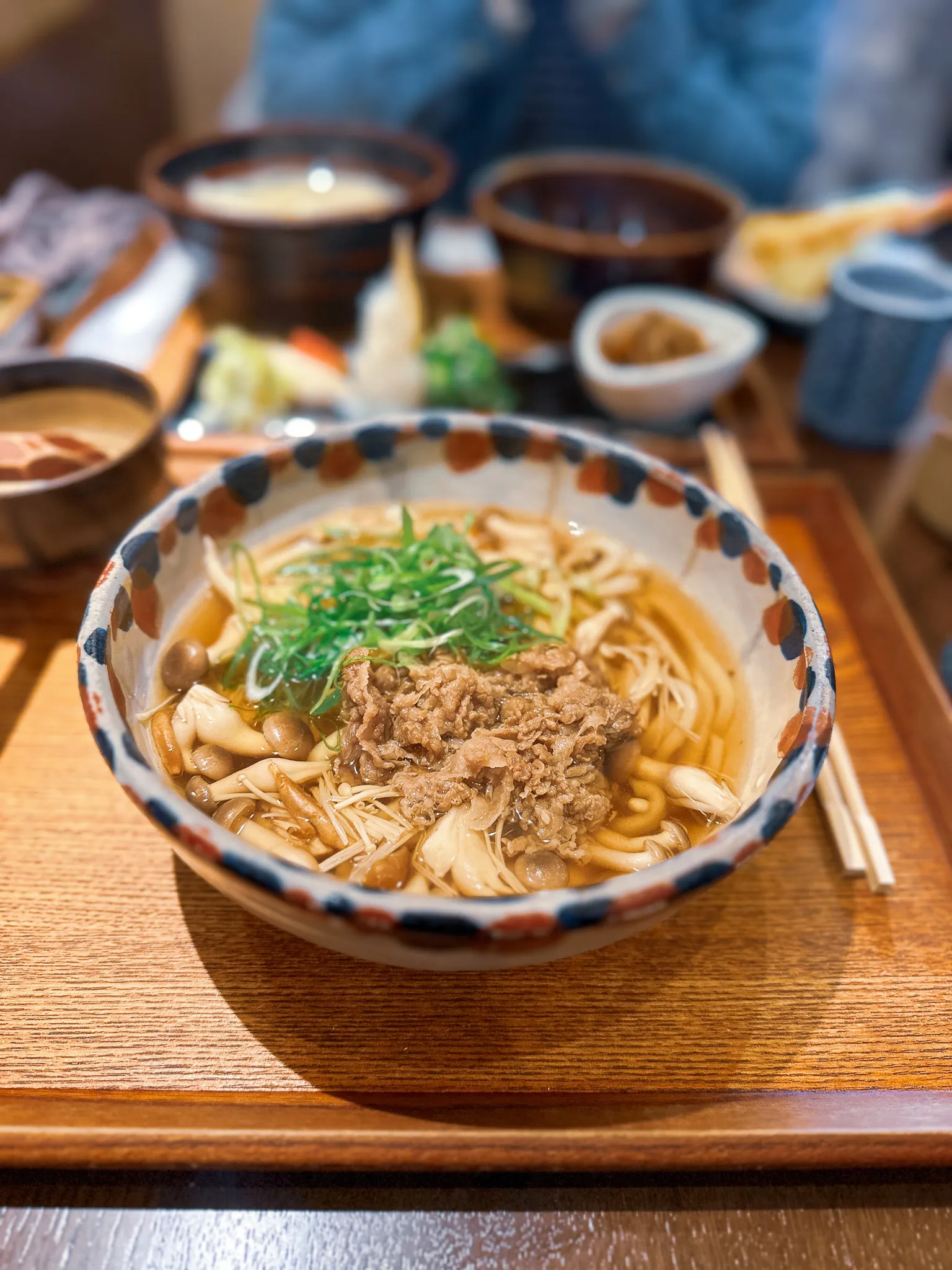 A bowl of udon with sliced beef, spring onions and several kinds of mushroom in a clear brown broth, on a wooden tray with chopsticks beside it