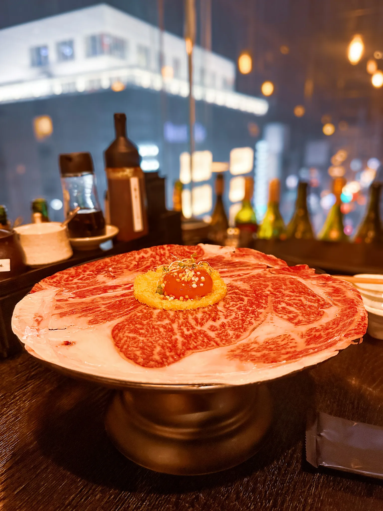 A wide plate of raw, heavily marbled wagyu on a brass pedestal stand, a crisp round and an egg yolk set in the middle, the street lights outside the window blurred behind