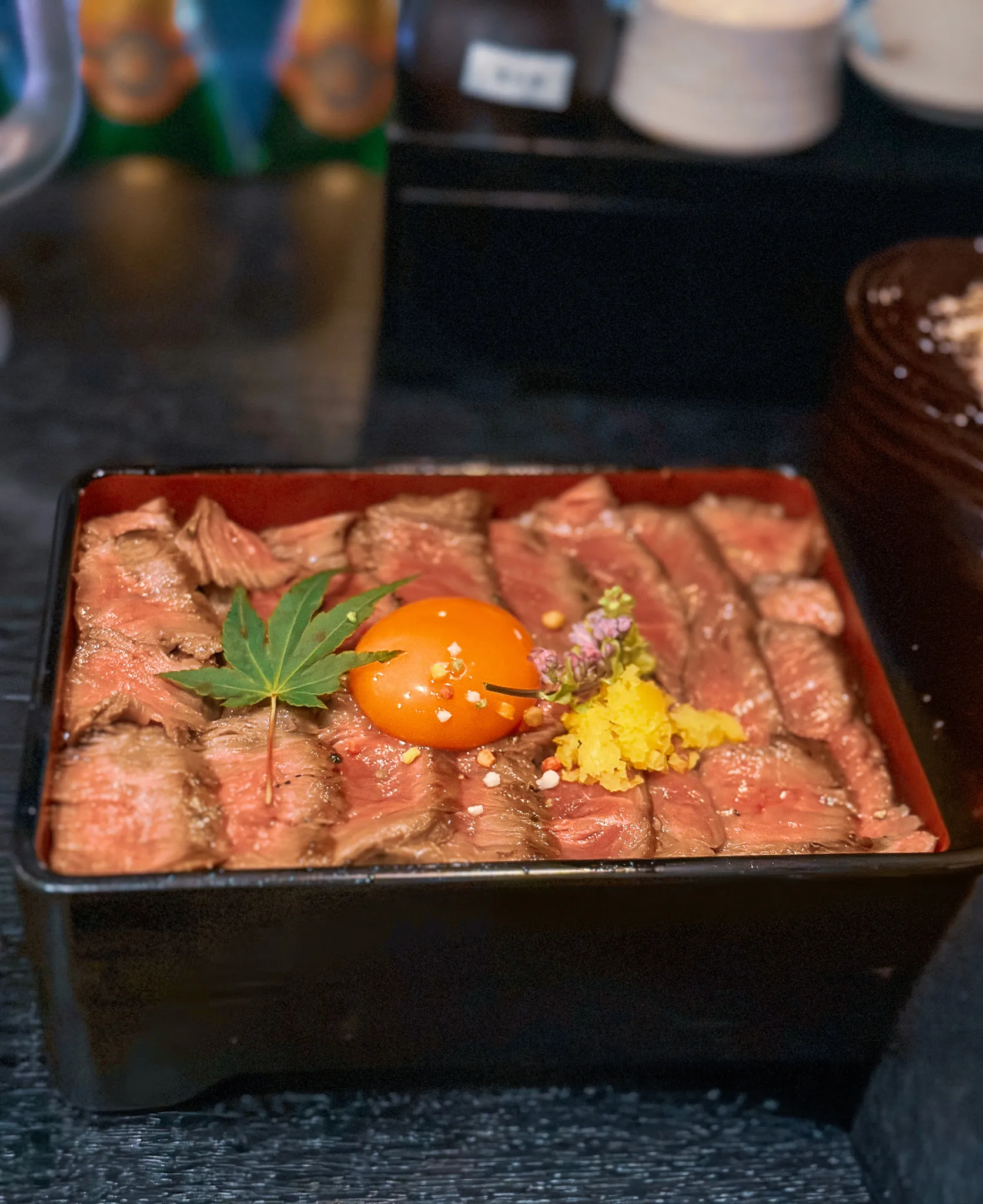 A lacquer box packed with slices of rare wagyu over rice, a whole egg yolk in the middle with a maple leaf, grated yuzu and a small flower beside it