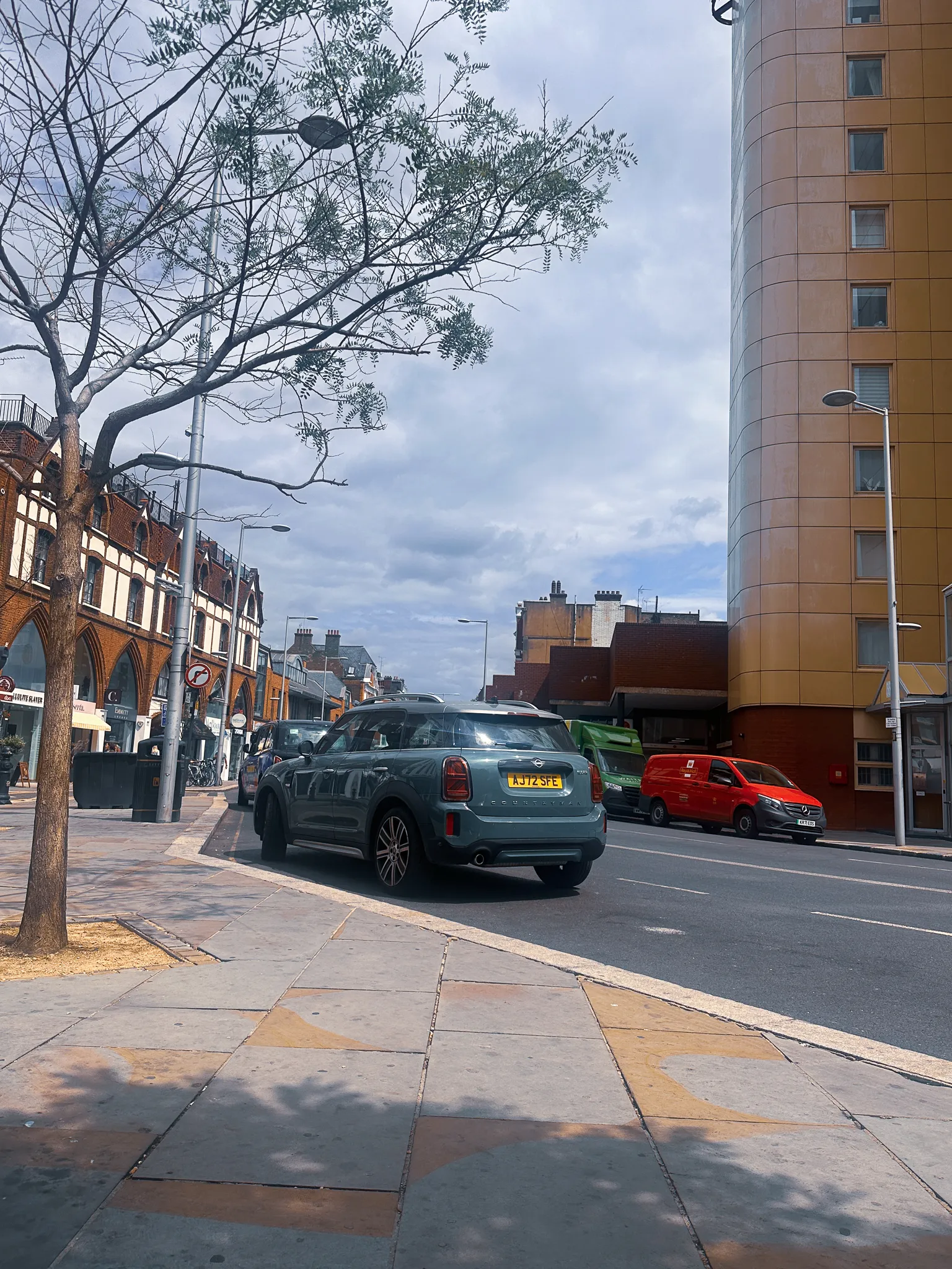 A west London high street on the first afternoon - a green Mini parked at the kerb, a red van and a green lorry waiting behind it, a brick parade of shops with pointed arches on the left and a tower block clad in yellow tile curving up on the right