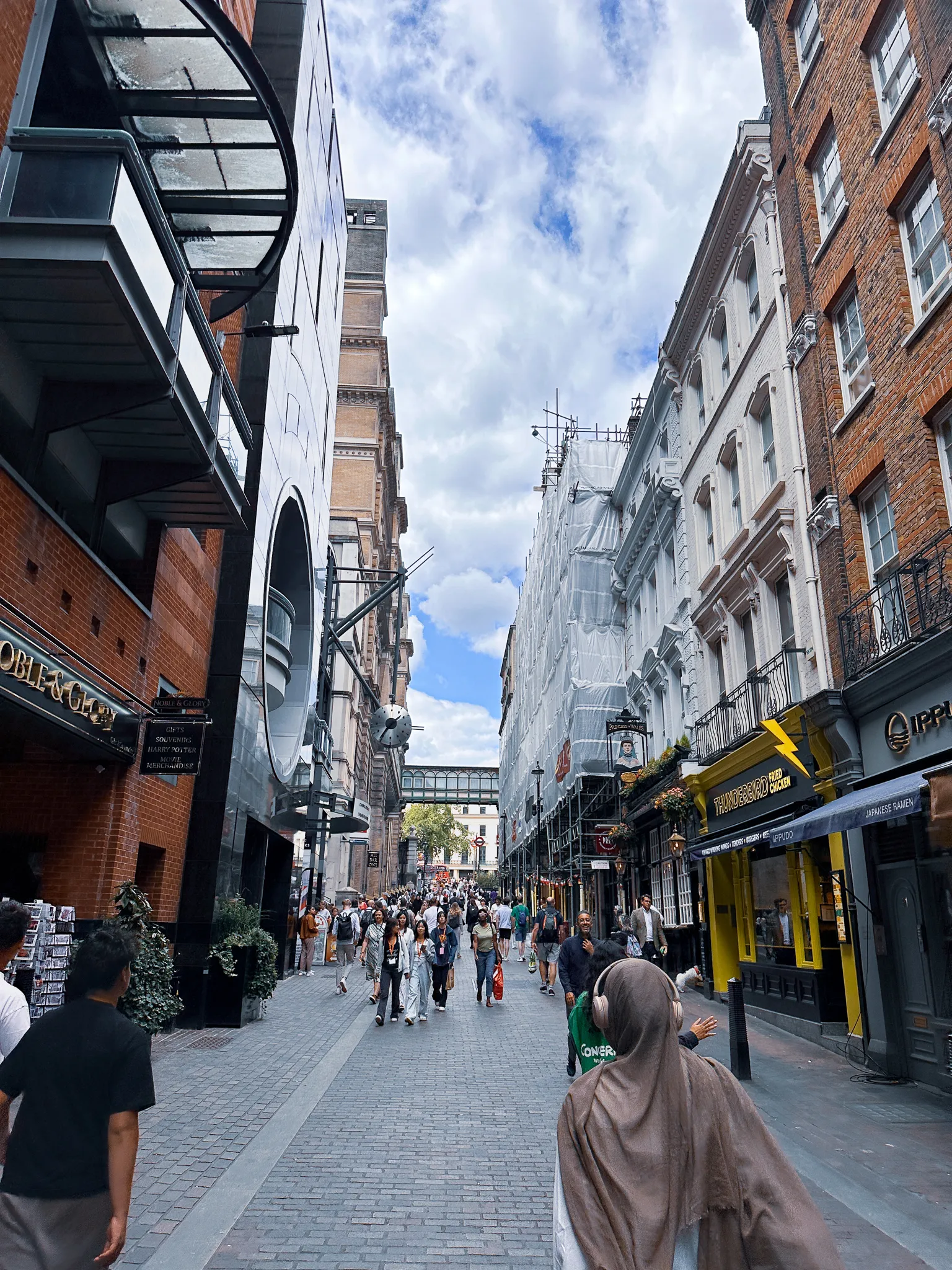 A narrow pedestrian street packed with people, a souvenir shop and a Japanese ramen bar on the left, a yellow fried chicken shopfront on the right and the glass arch of a station roof closing the far end
