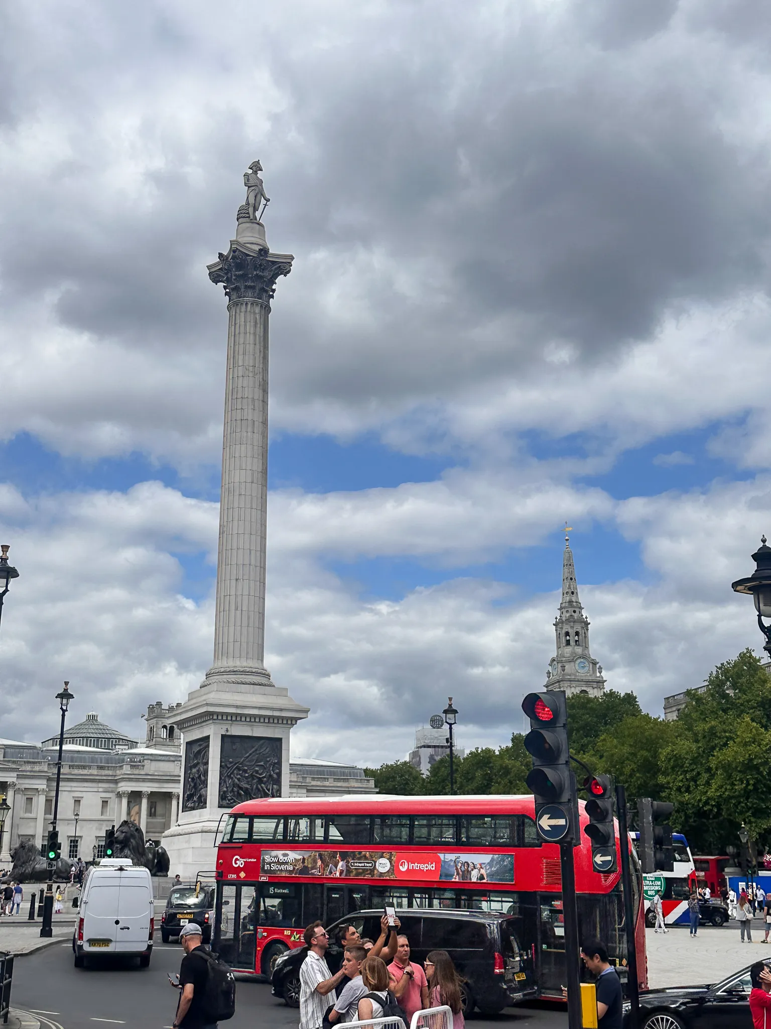 Nelson's Column under heavy grey cloud, one of the bronze lions on its plinth at the left, the spire of St Martin-in-the-Fields to the right and a red bus crossing the front of the square