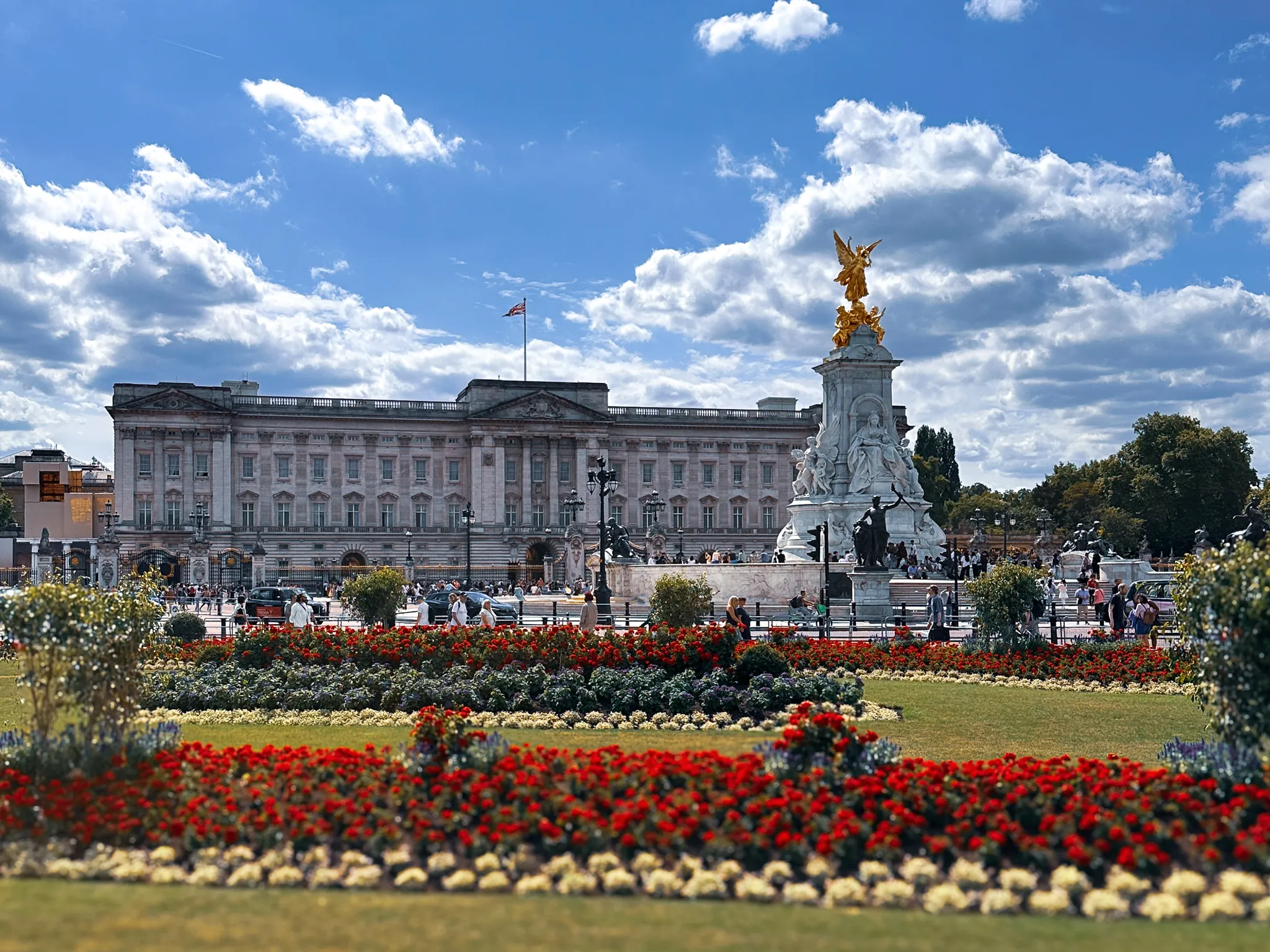 Buckingham Palace across the memorial gardens, the gilded Victory on the Victoria Memorial catching the sun, beds of red and white flowers filling the foreground and the flag flying over the roof