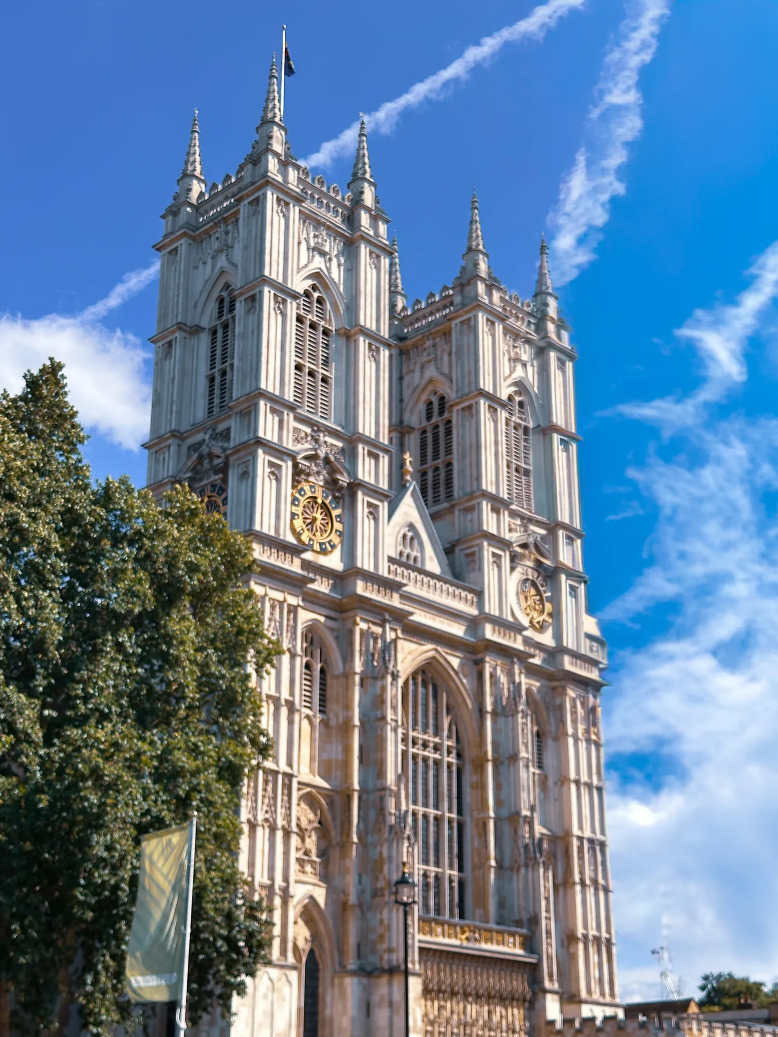 The west towers of Westminster Abbey from below, the blue and gold clock face on the left tower, contrails drawn across a deep blue sky and a plane tree at the edge of the frame