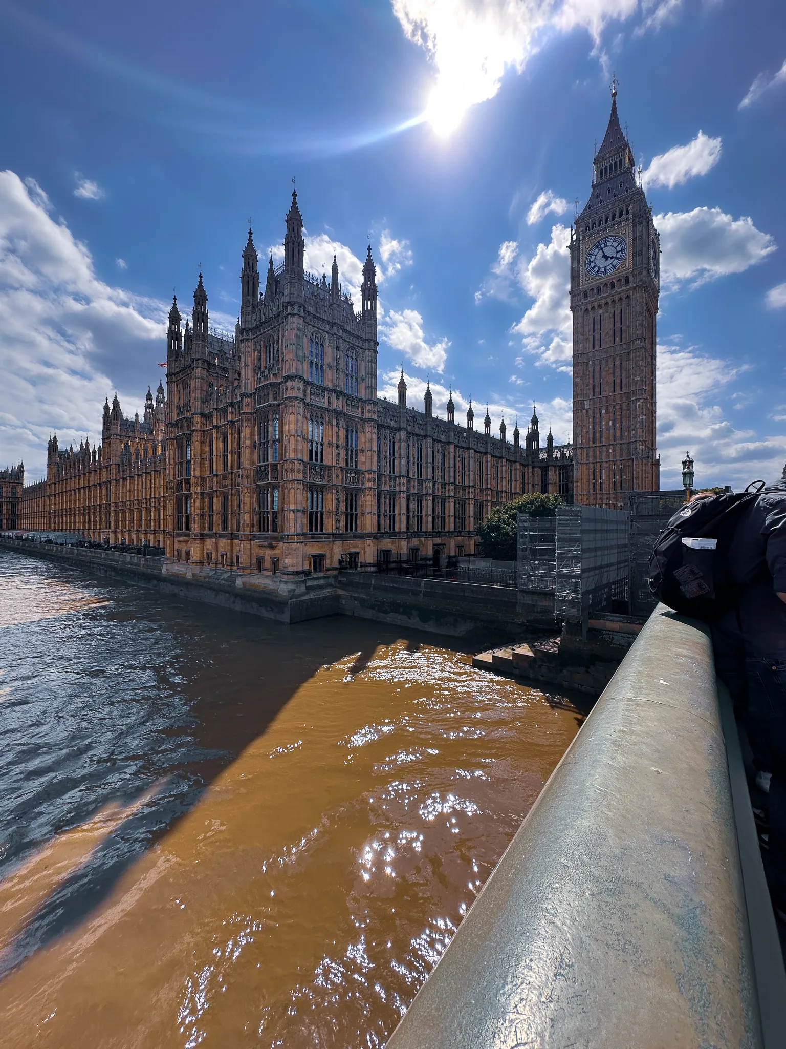 The Palace of Westminster from Westminster Bridge with the sun flaring above the Elizabeth Tower, the brown Thames running below and somebody's backpack resting on the bridge rail in the corner