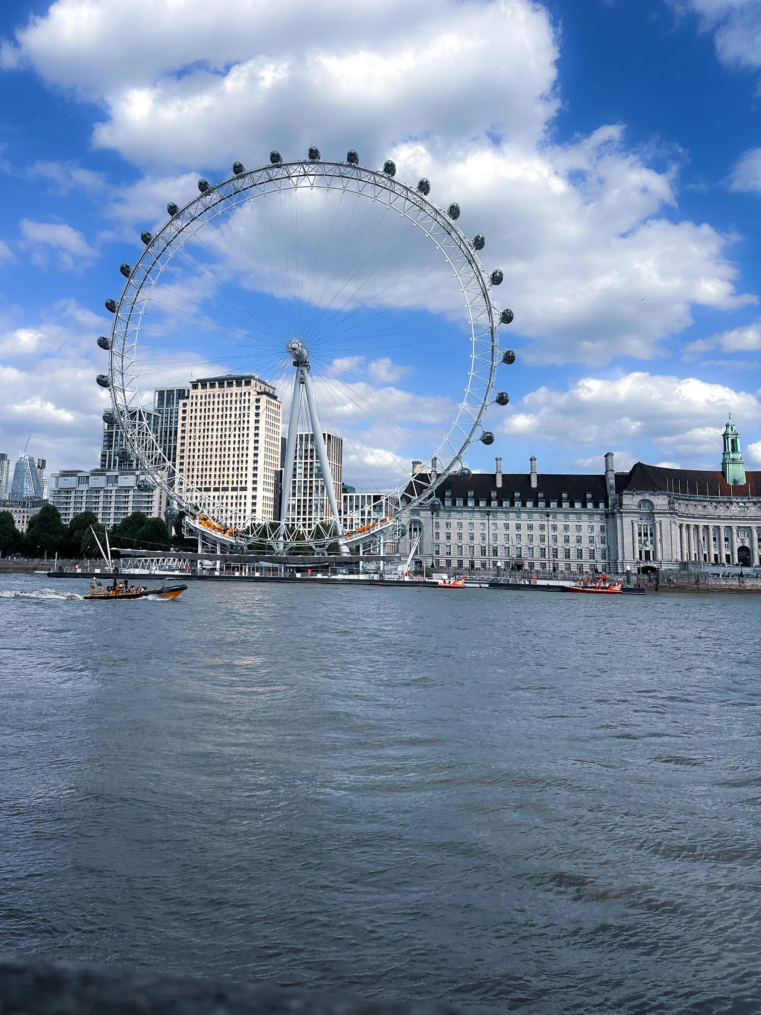 The London Eye across the Thames with County Hall beside it, a speedboat throwing a wake through grey water and clouds piling up behind the wheel