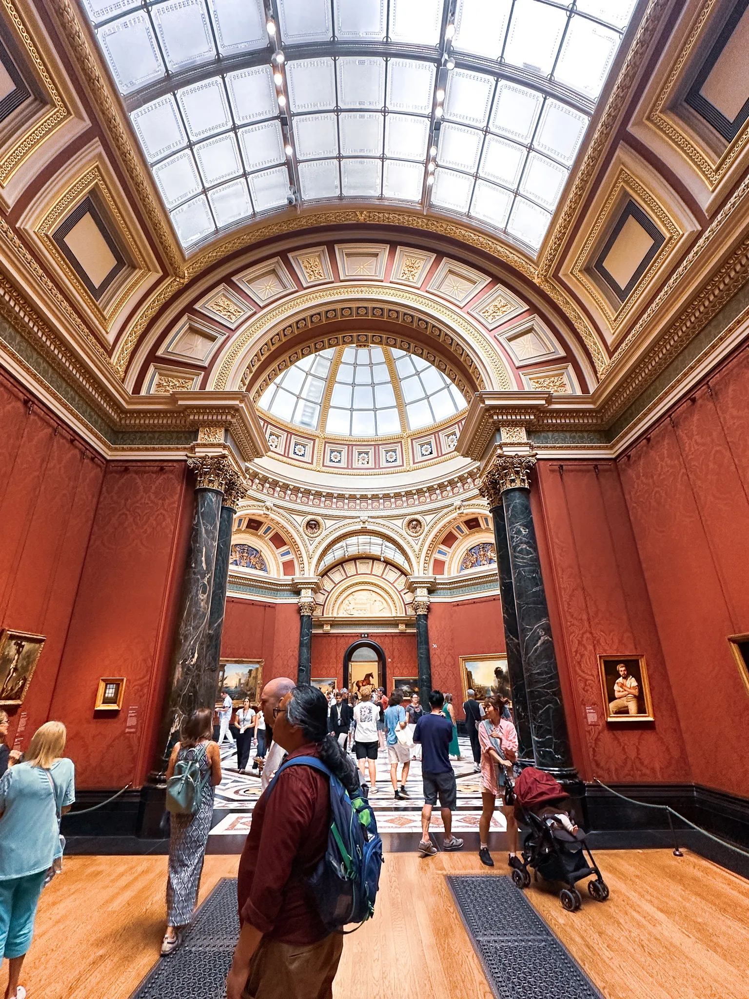 Inside the National Gallery - deep red walls hung with paintings, green marble columns either side, a glass dome and barrel roof overhead and visitors crossing the mosaic floor