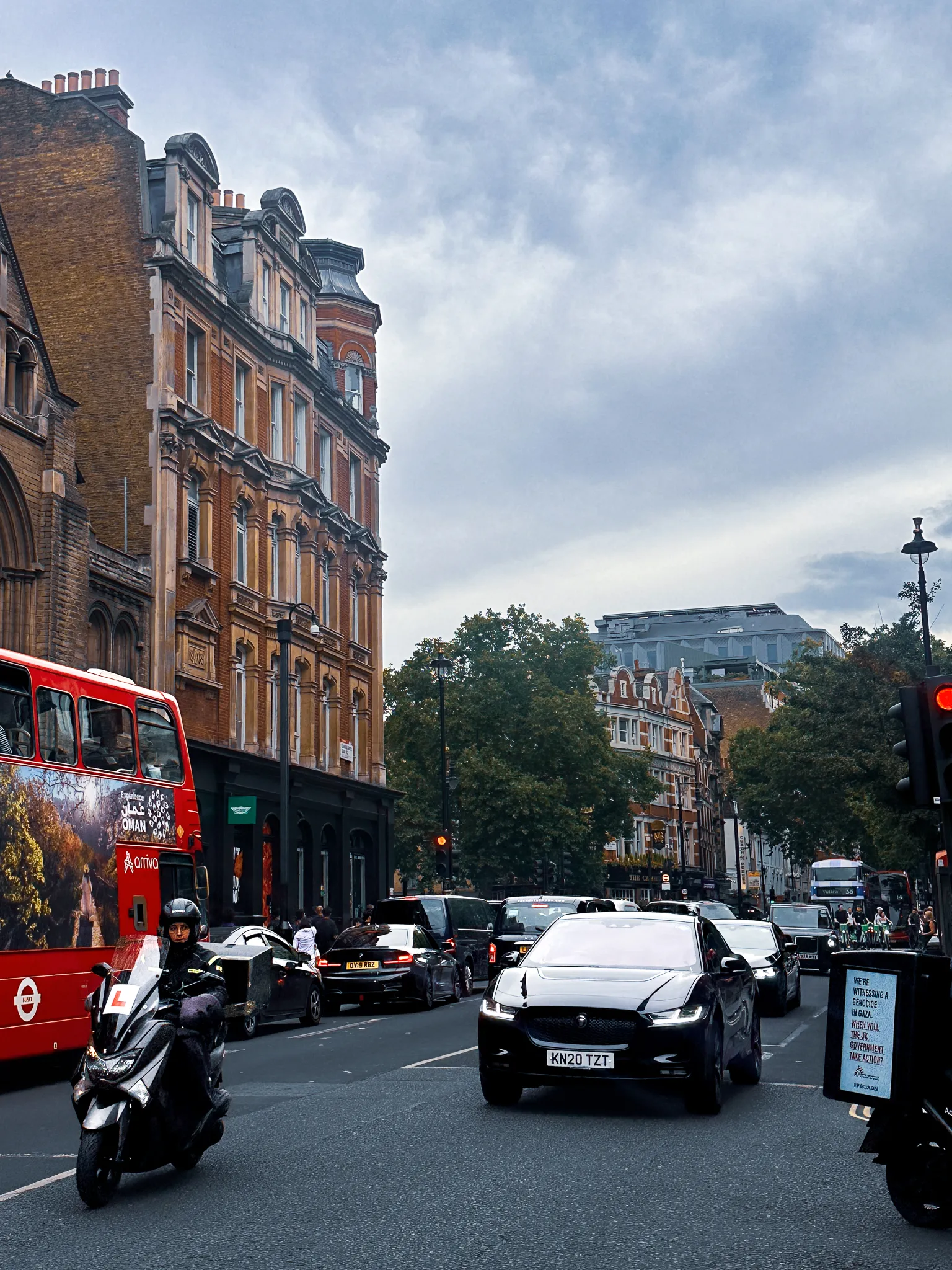 Evening traffic under a flat grey sky, a learner on a scooter alongside a red bus, an electric Jaguar in the middle of the road and Victorian brick and terracotta running up the left