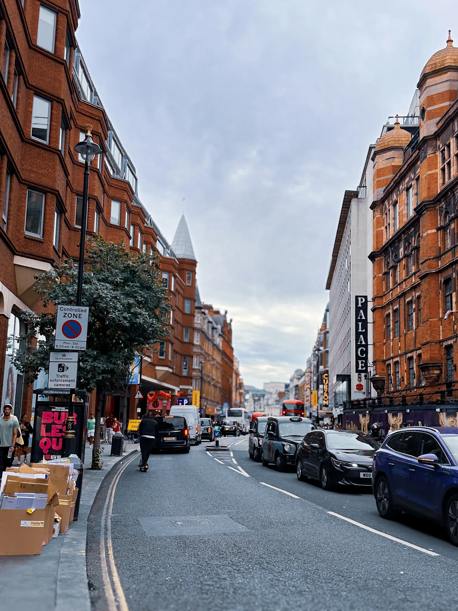 Shaftesbury Avenue in the evening, the Palace Theatre's vertical sign lit on the right, red brick mansion blocks down both sides, flattened cardboard stacked at the kerb and buses queued in the distance