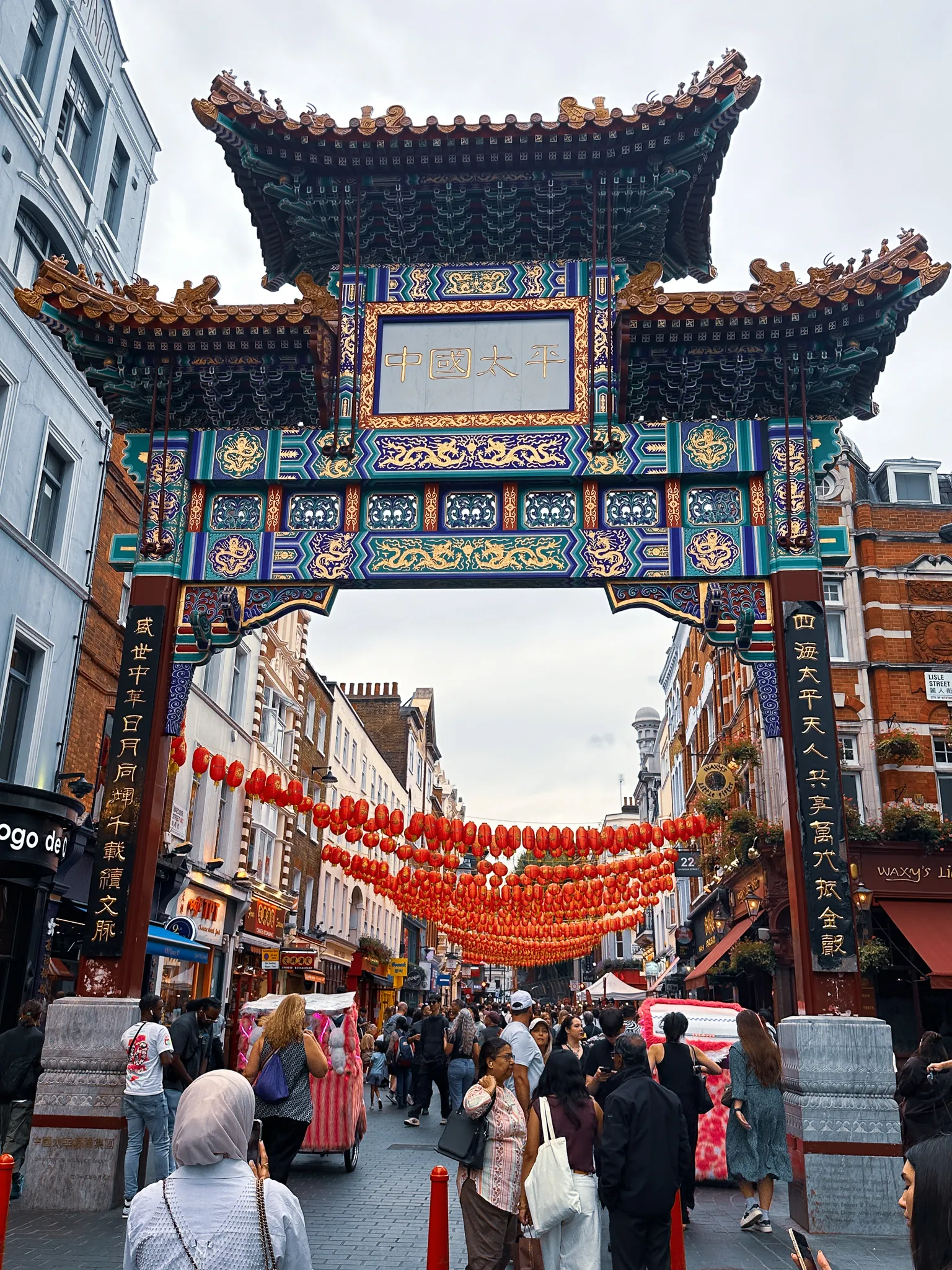 The tall painted archway into Chinatown, green and gold with a blue panel of characters, ropes of red and orange lanterns hanging down the street beyond it and a crowd passing underneath