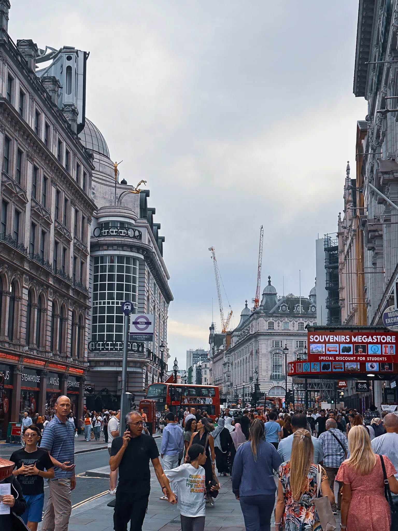 The crowd on Coventry Street heading for Piccadilly Circus, a souvenir megastore's red sign on the right, cranes over the rooftops and a red bus stopped at the junction ahead