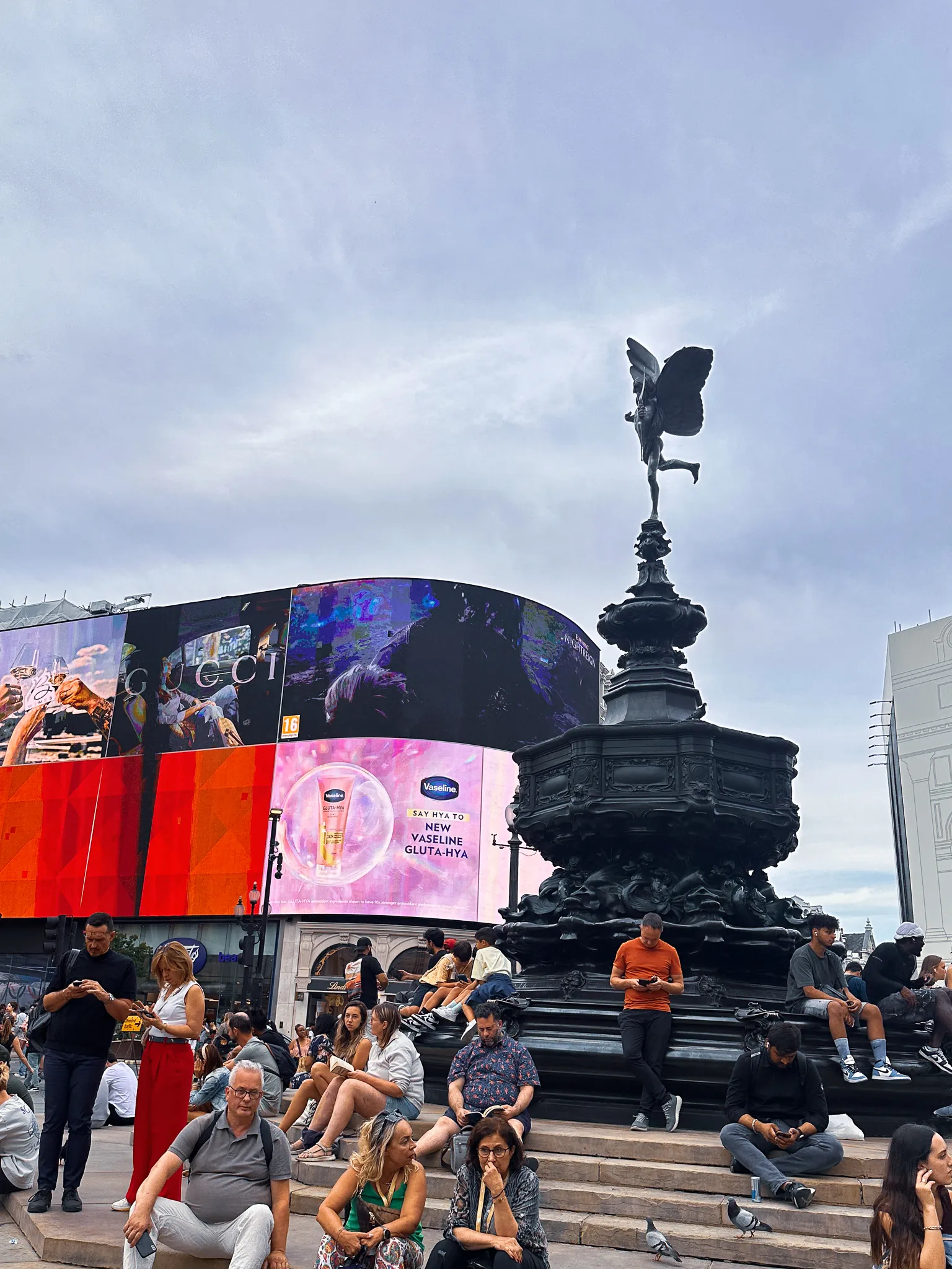 The winged archer on the Shaftesbury Memorial above the fountain at Piccadilly Circus, the curved advertising screens lit behind it and people sitting all down the steps