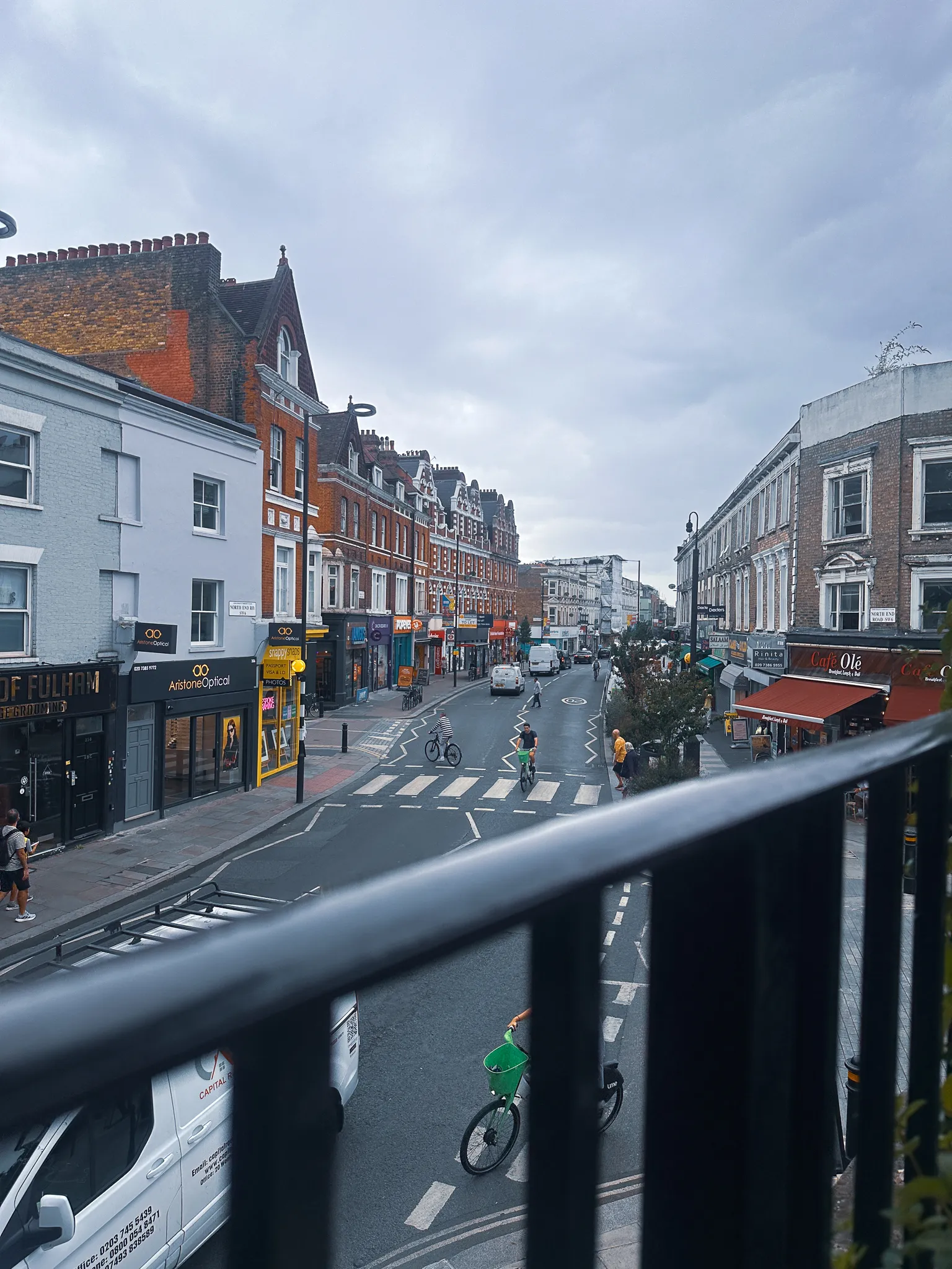 A Fulham street from a first-floor balcony - shopfronts and a café's red awning, a zebra crossing, cyclists waiting at the junction and a black railing across the foreground