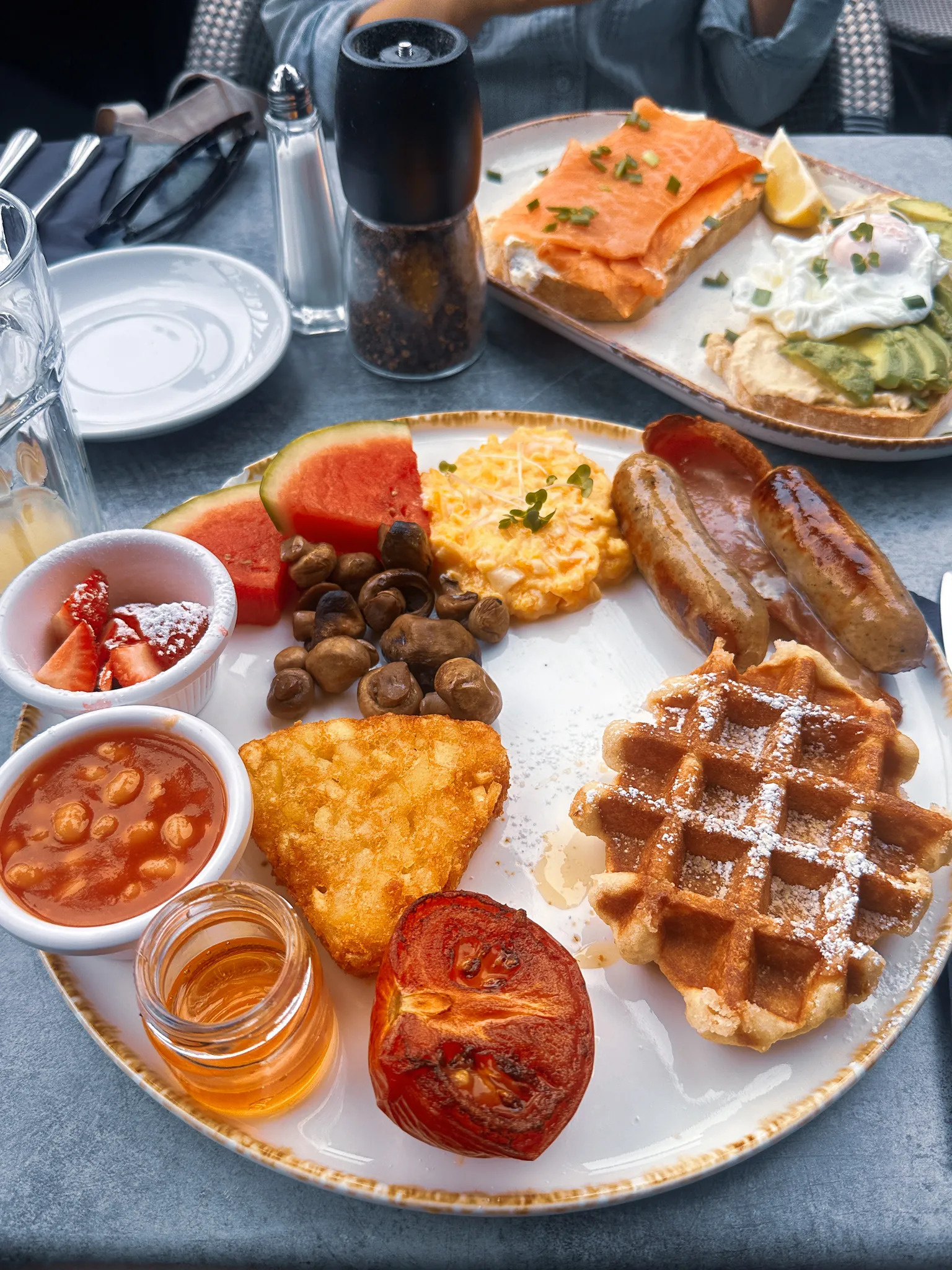 A brunch plate loaded with scrambled egg, sausages, bacon, a hash brown, mushrooms, a grilled tomato and a sugared waffle, ramekins of beans and strawberries beside it, with smoked salmon and avocado toasts on the plate behind