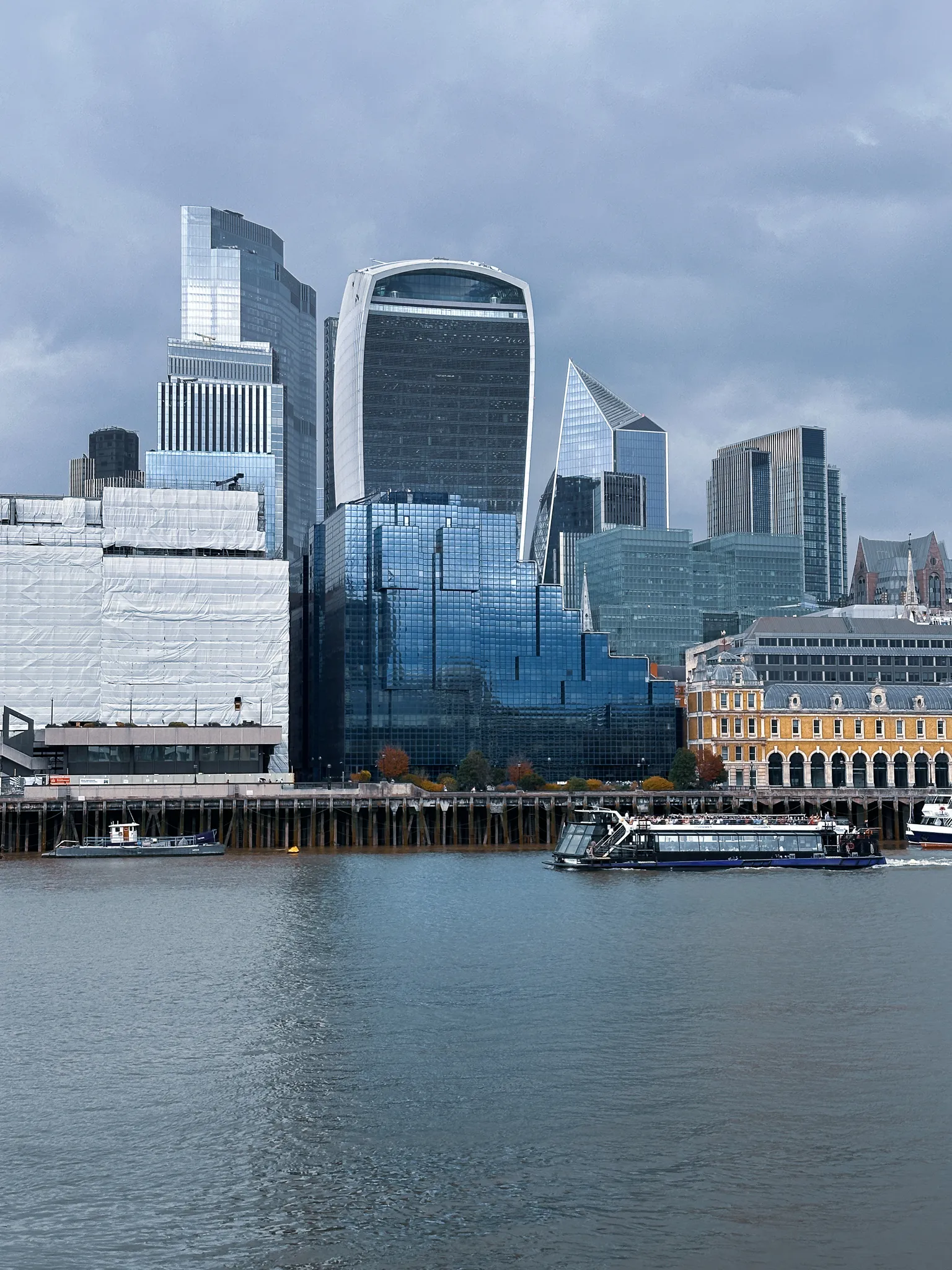 The City across the river under grey cloud - the Walkie-Talkie in the middle, a wedge-shaped tower behind it, a low yellow-brick market building at the water's edge and a tour boat passing