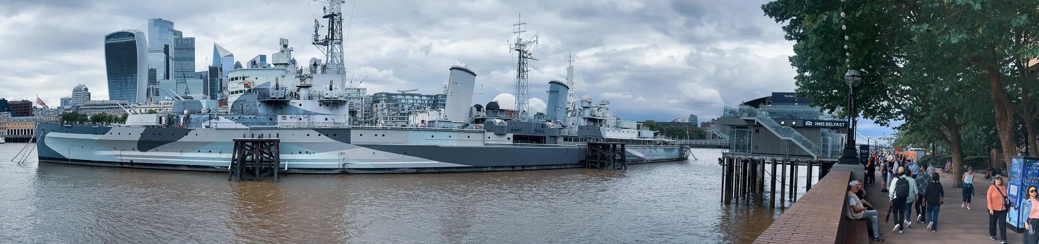 HMS Belfast moored on the south bank in her grey dazzle paint, the towers of the City rising behind her and the riverside walk under plane trees on the right