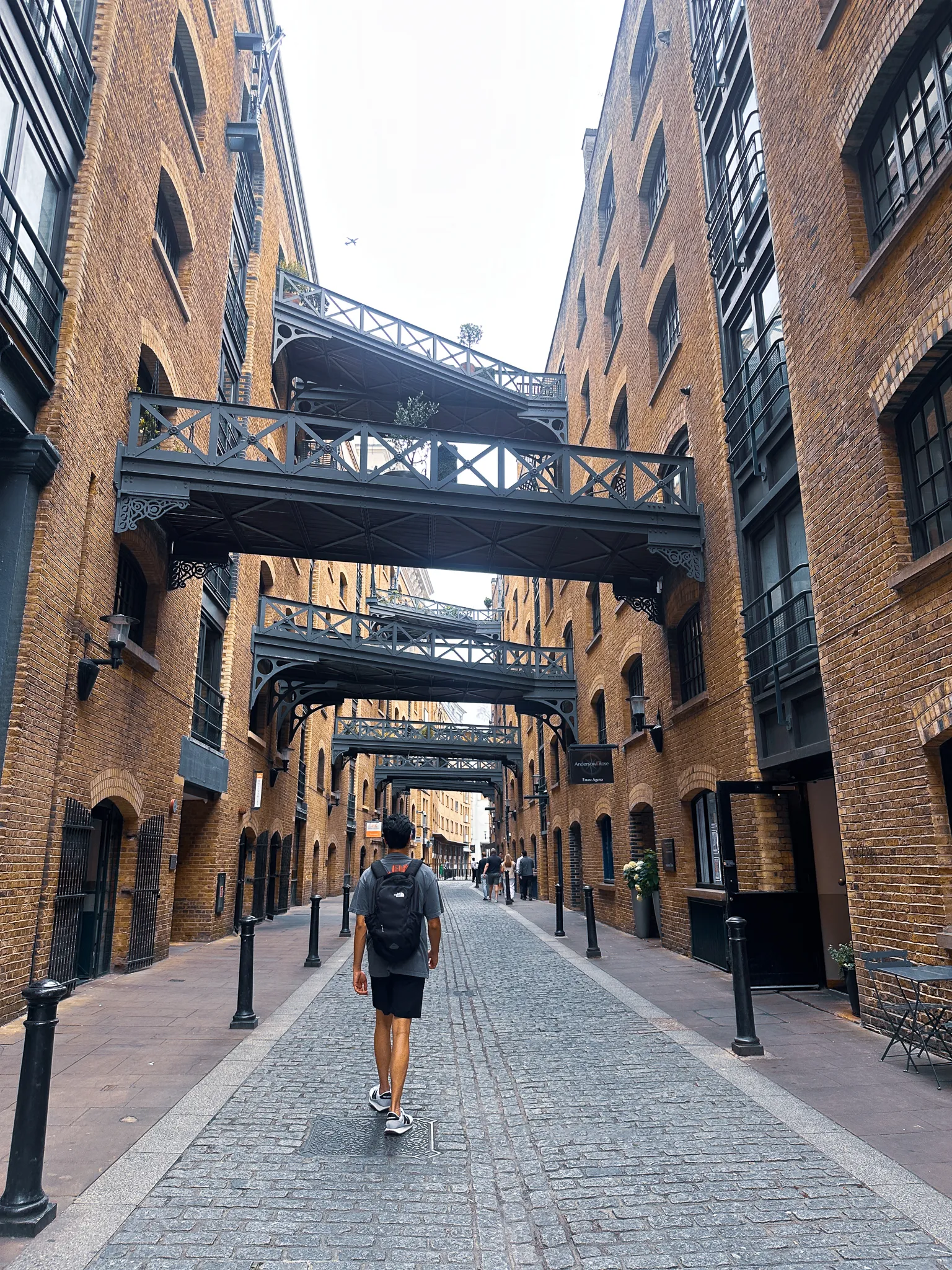 The warehouse canyon at Shad Thames - iron footbridges crossing overhead between brick wharves, cobbles underfoot and a man walking away down the middle of it