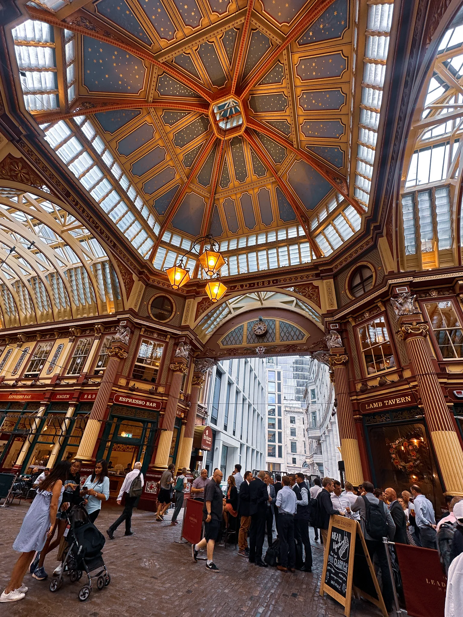Leadenhall Market under its painted iron roof, dark red and gold with stars on the ceiling, lanterns hanging at the crossing and drinkers in suits standing outside the Lamb Tavern