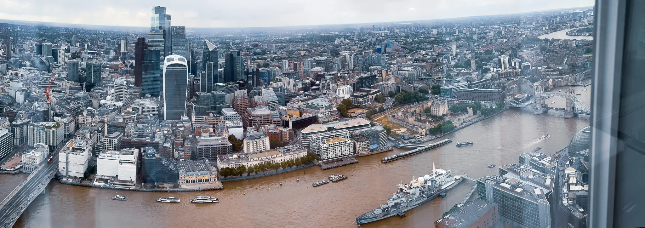North from high above the river - the towers of the City on the left, the Thames curving through the middle with Tower Bridge and the Tower of London beyond and HMS Belfast tied up below