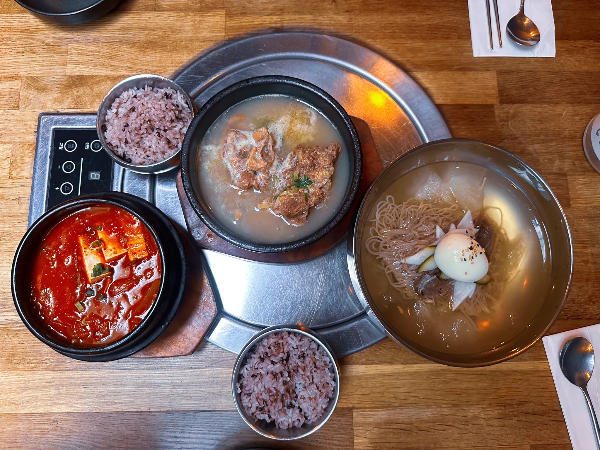 A Korean dinner from above on a steel table - a stone pot of milky bone soup, a black pot of red kimchi stew, two bowls of purple multigrain rice and a wide steel bowl of cold noodles with radish, cucumber and half an egg