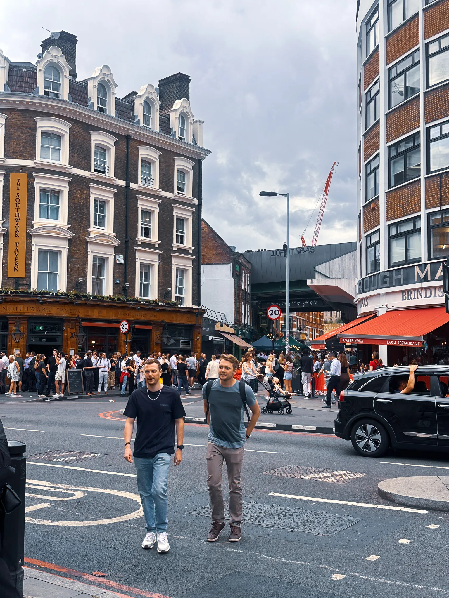 The corner by Borough Market at the end of the day, the Southwark Tavern's yellow sign above a crowd drinking on the pavement, red awnings across the road and two men crossing towards the camera
