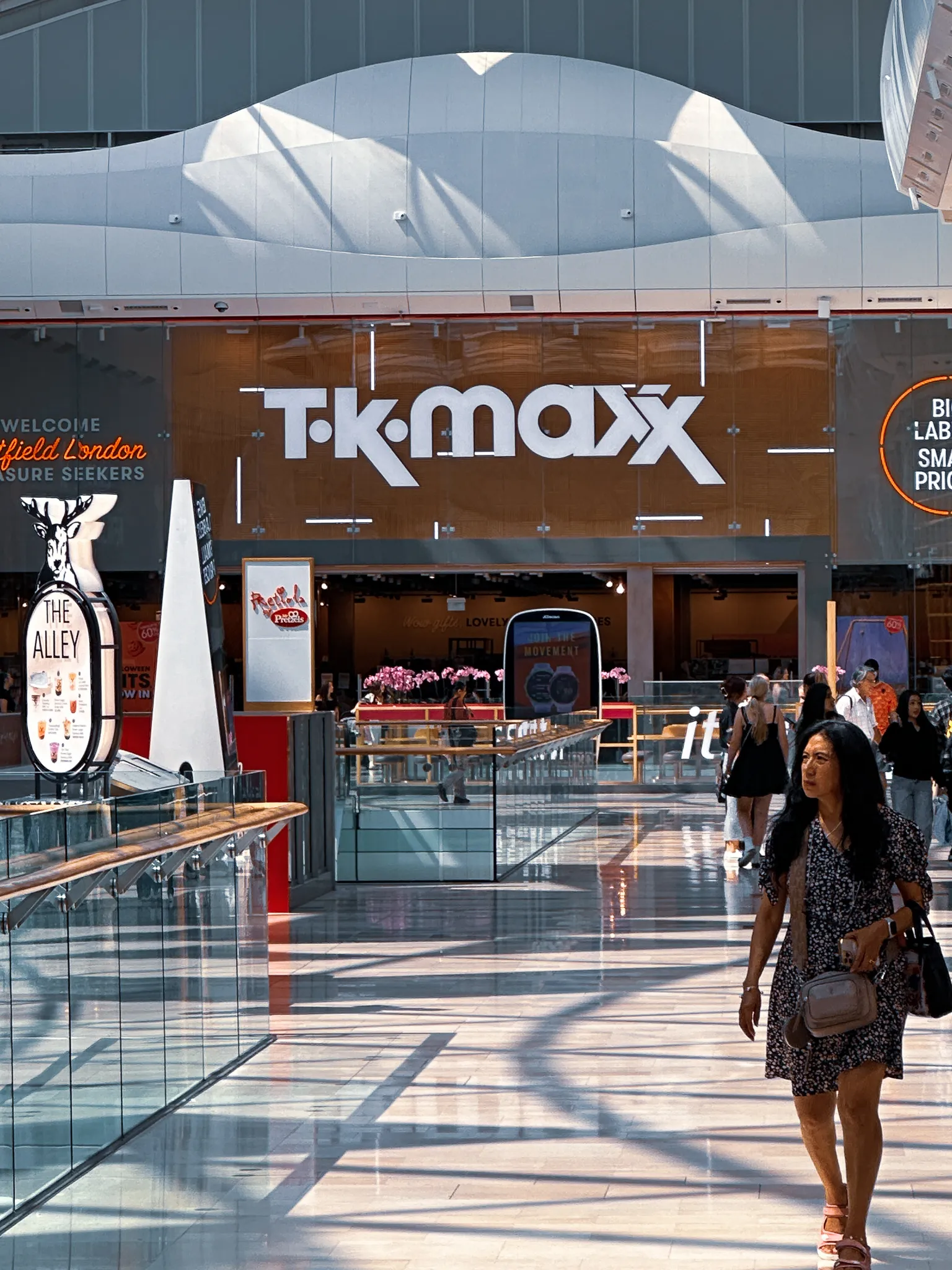 Inside Westfield London - a TK Maxx storefront under a white wave of a roof, a bubble tea sign on the left and the polished floor striped with shadow from the glass above