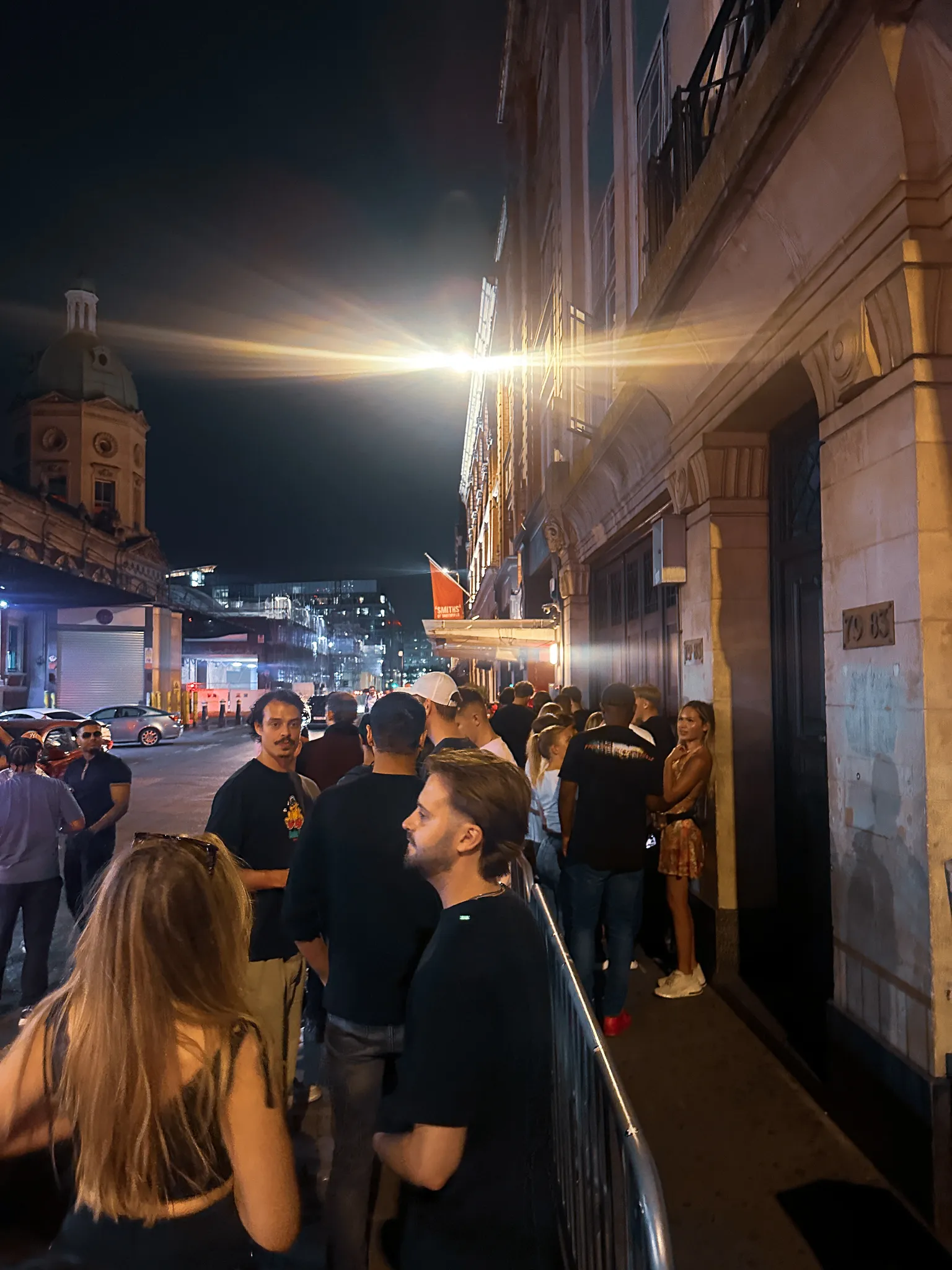 A queue along the pavement after midnight, barriers down one side, a streetlight flaring overhead and the dome of an old building at the end of the street