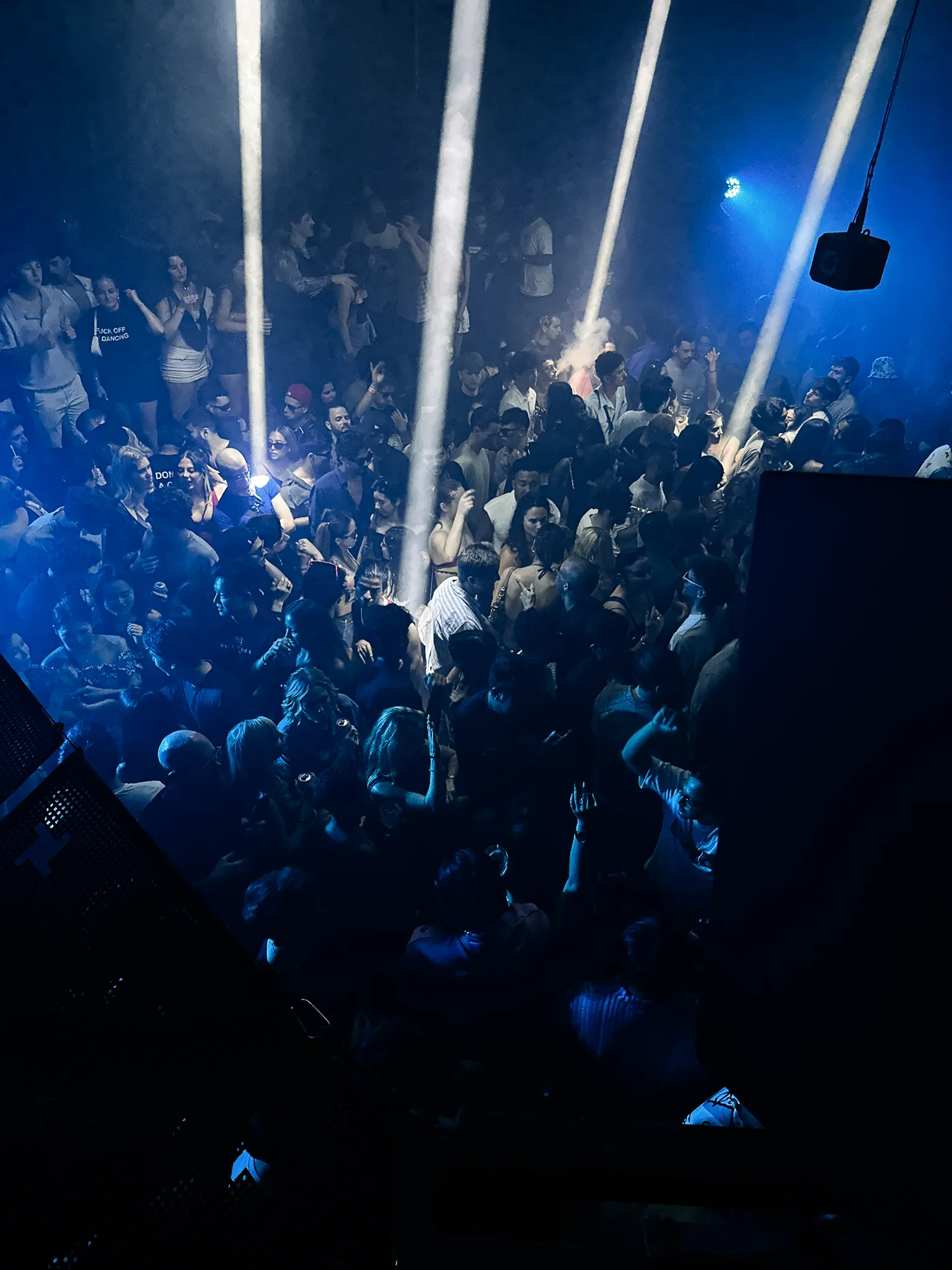 The dancefloor from above, beams of white light cutting through haze over a packed crowd, a speaker hanging into the frame and the whole room washed blue