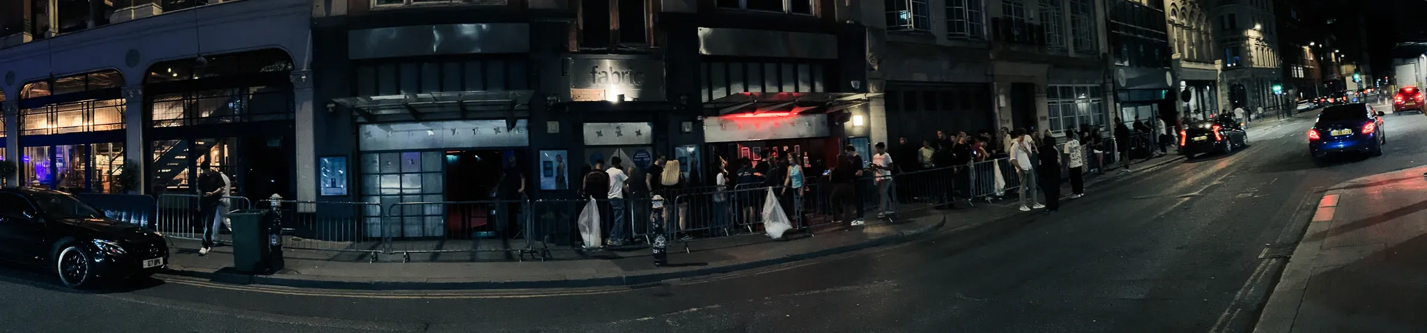 A wide view of the corner outside the club - the line of people along the barriers, the red-lit doorway, a car waiting at the kerb and the street dark behind