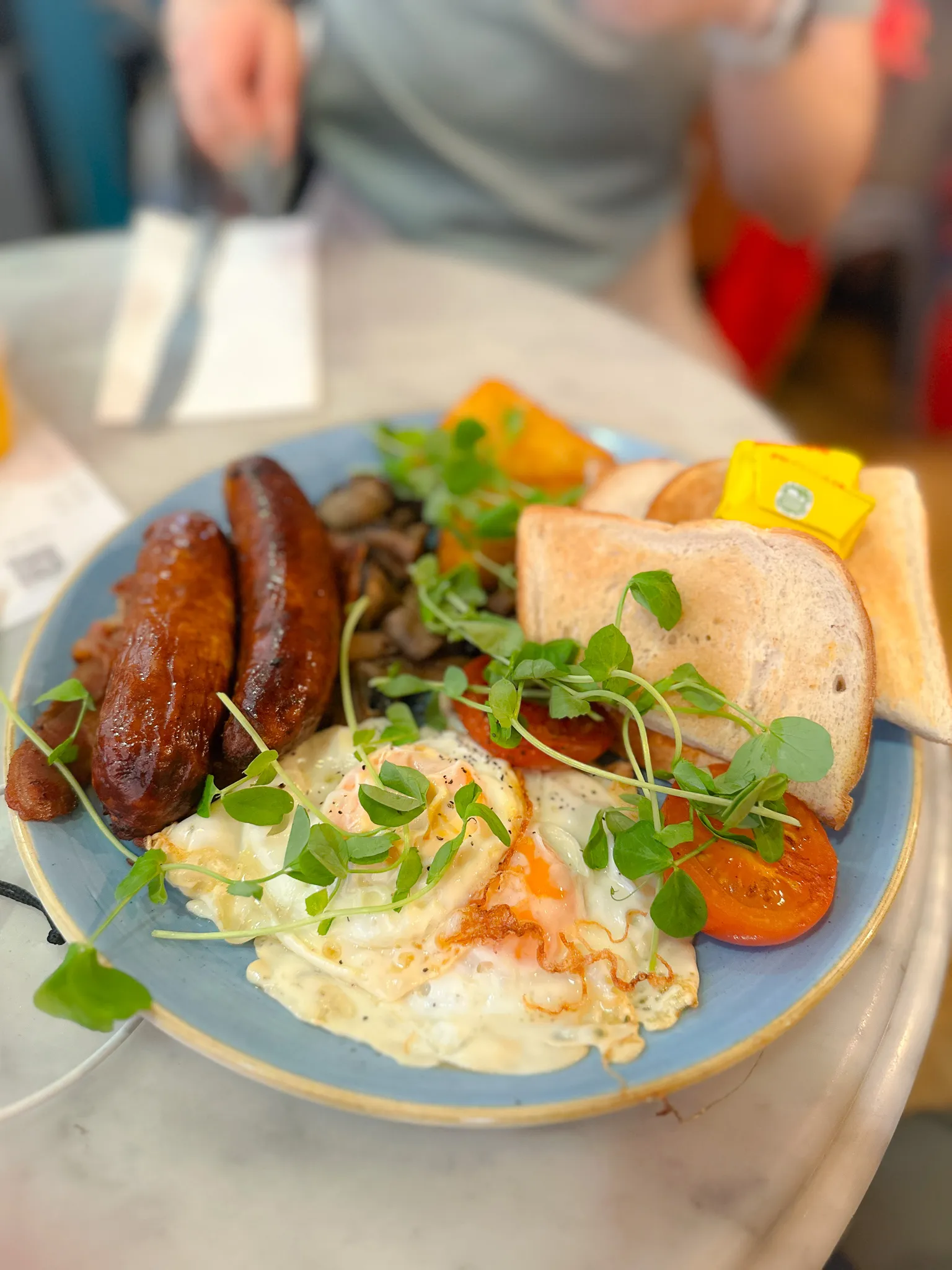 A full English on a blue plate - two sausages, bacon, fried eggs, mushrooms, a grilled tomato, a hash brown and toast with a pat of butter, pea shoots scattered over the top
