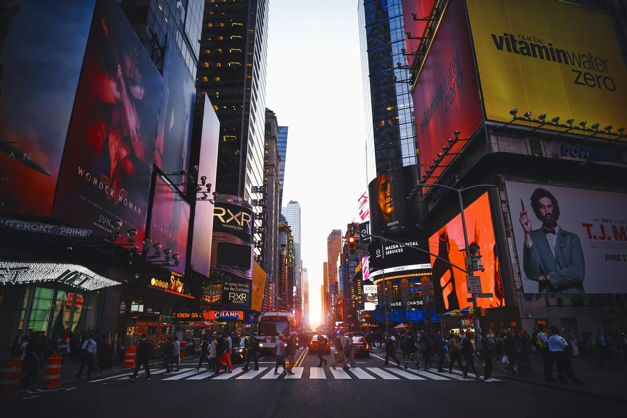 Times Square by day looking down the avenue into low sun, billboards on both sides and crowds spilling across the crossing