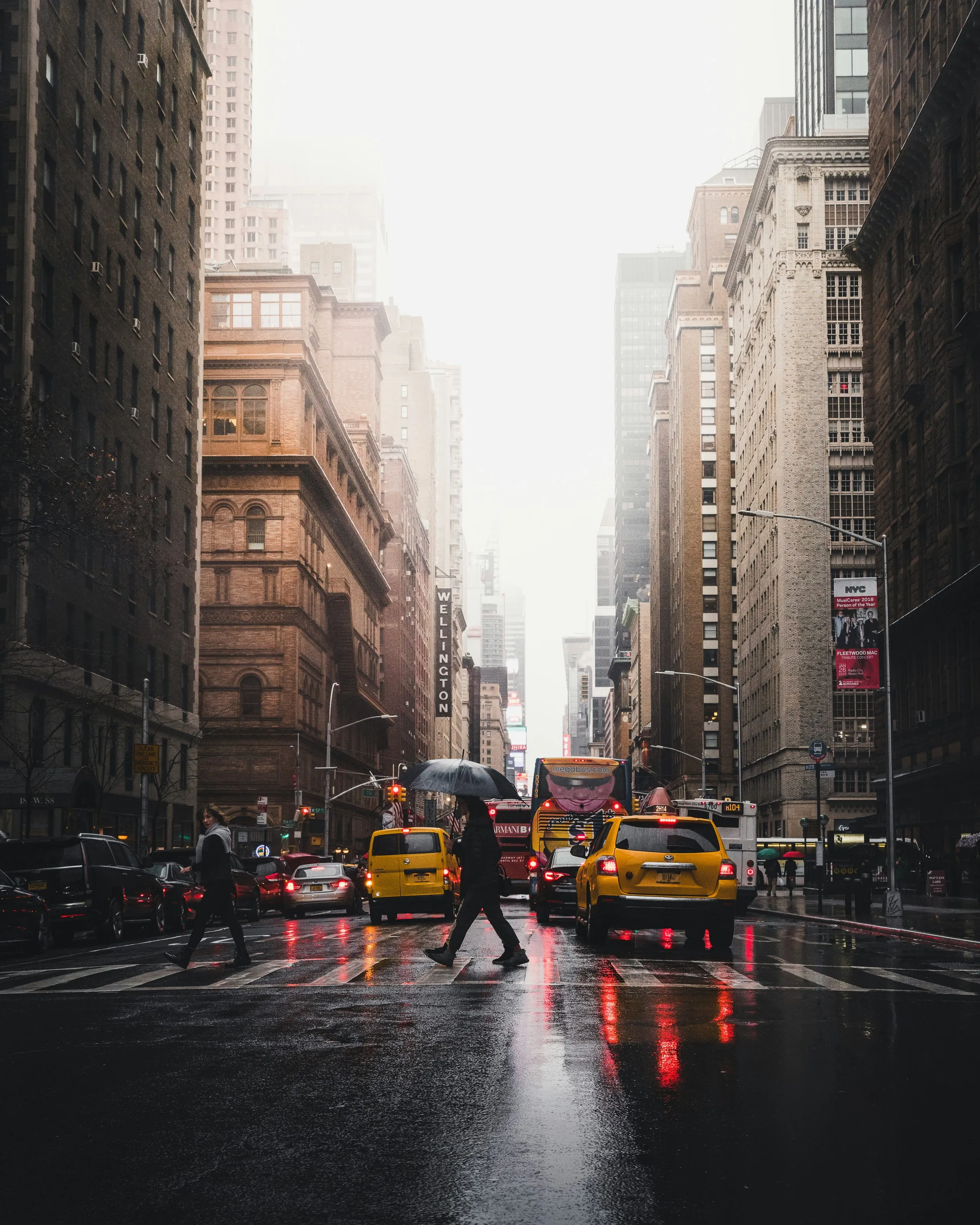 A wet street in the rain, people crossing under umbrellas past yellow cabs, tall buildings receding into white mist