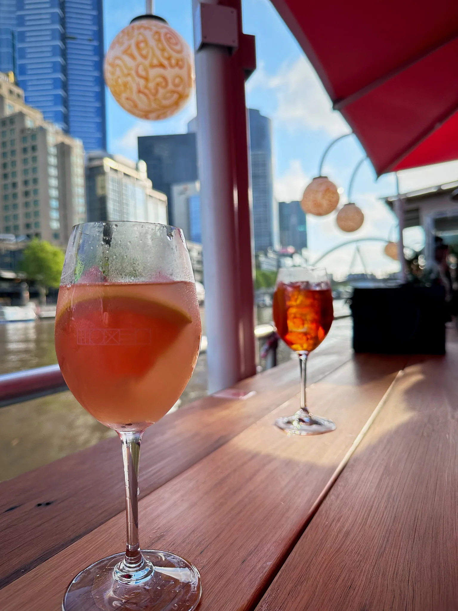 Two spritzes on a riverside bar table, paper lanterns and a red umbrella above, the Yarra behind