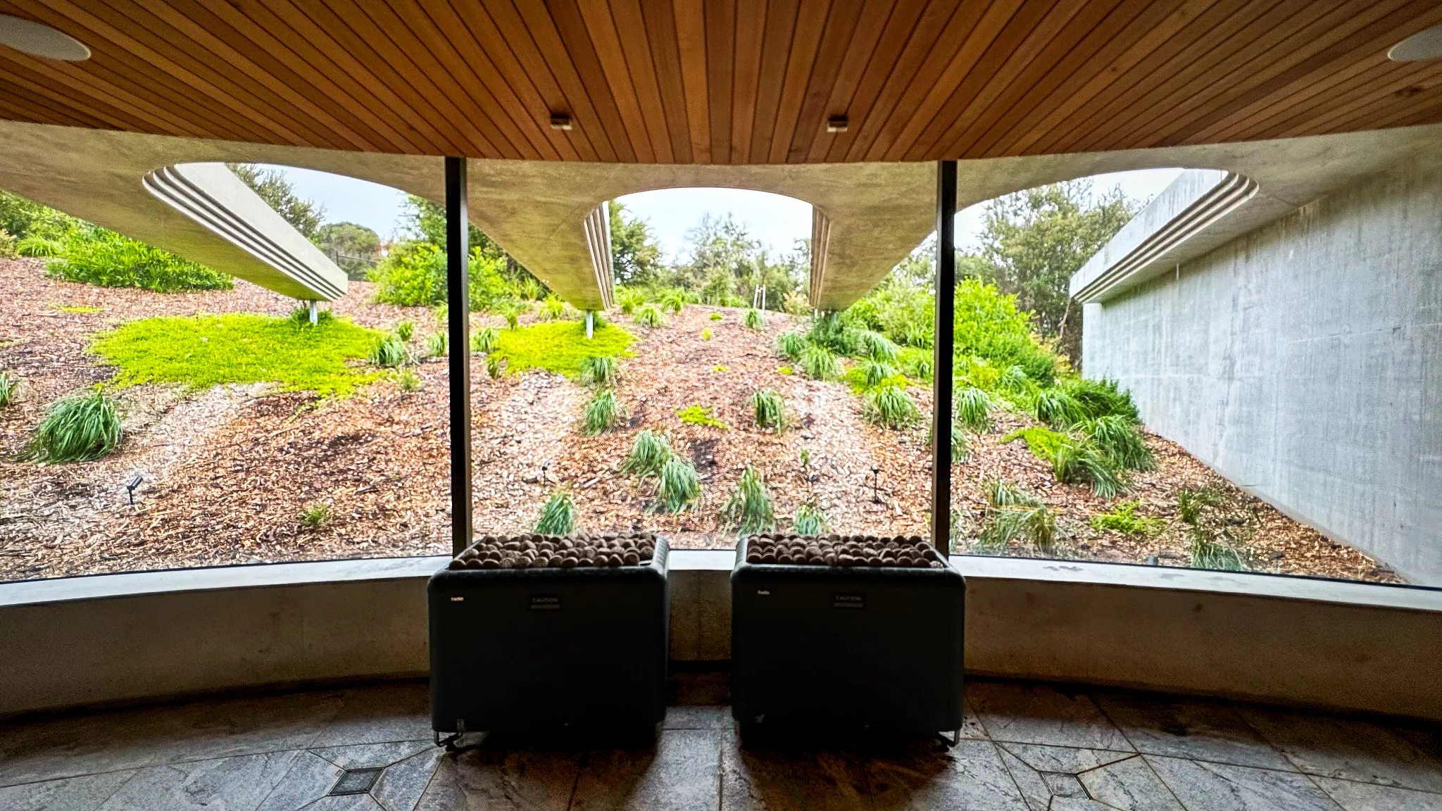 A curved window wall under a timber ceiling looking out at concrete arcs and planted slopes