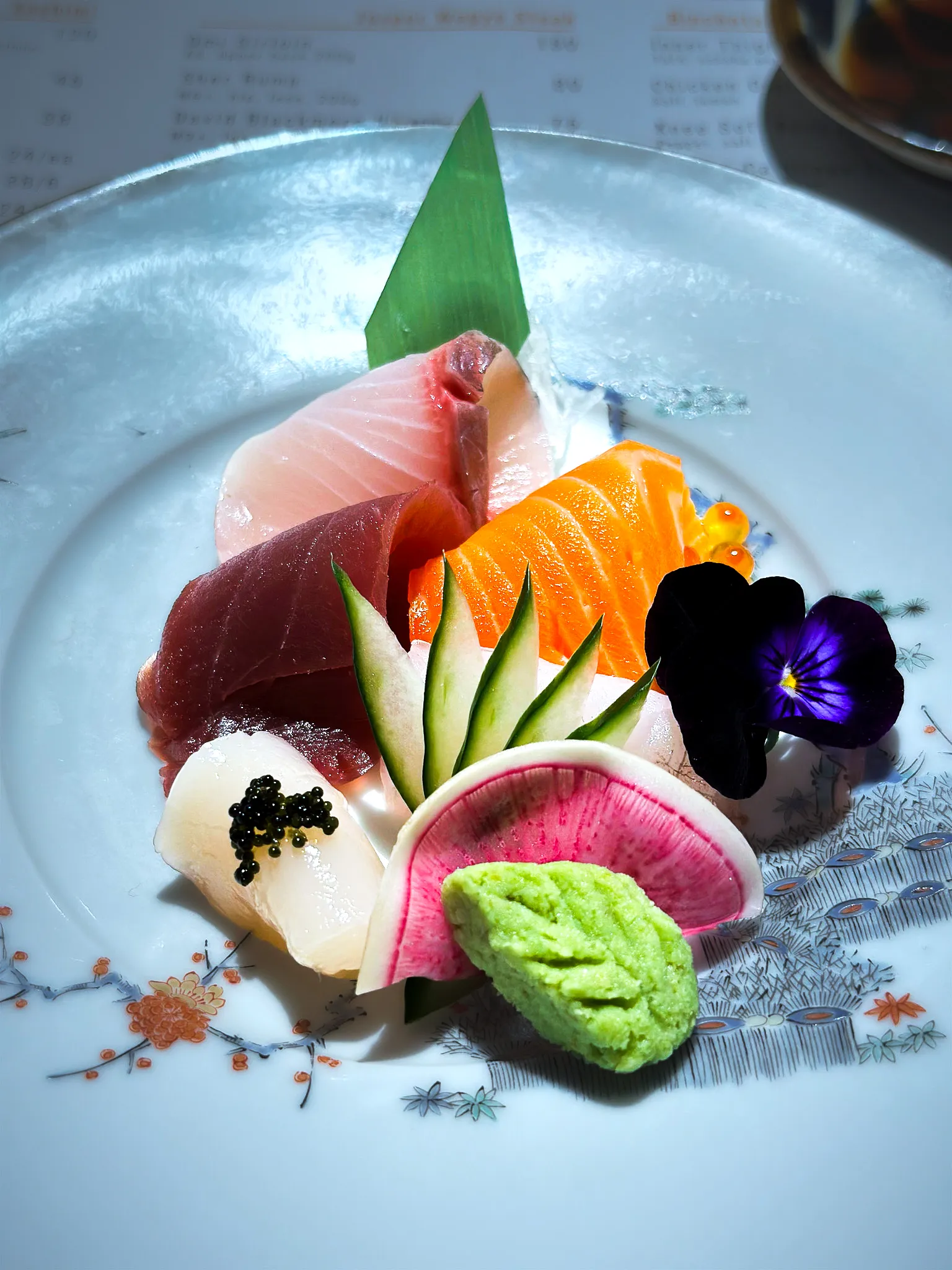 A plate of sashimi with cucumber, watermelon radish, wasabi and an edible pansy