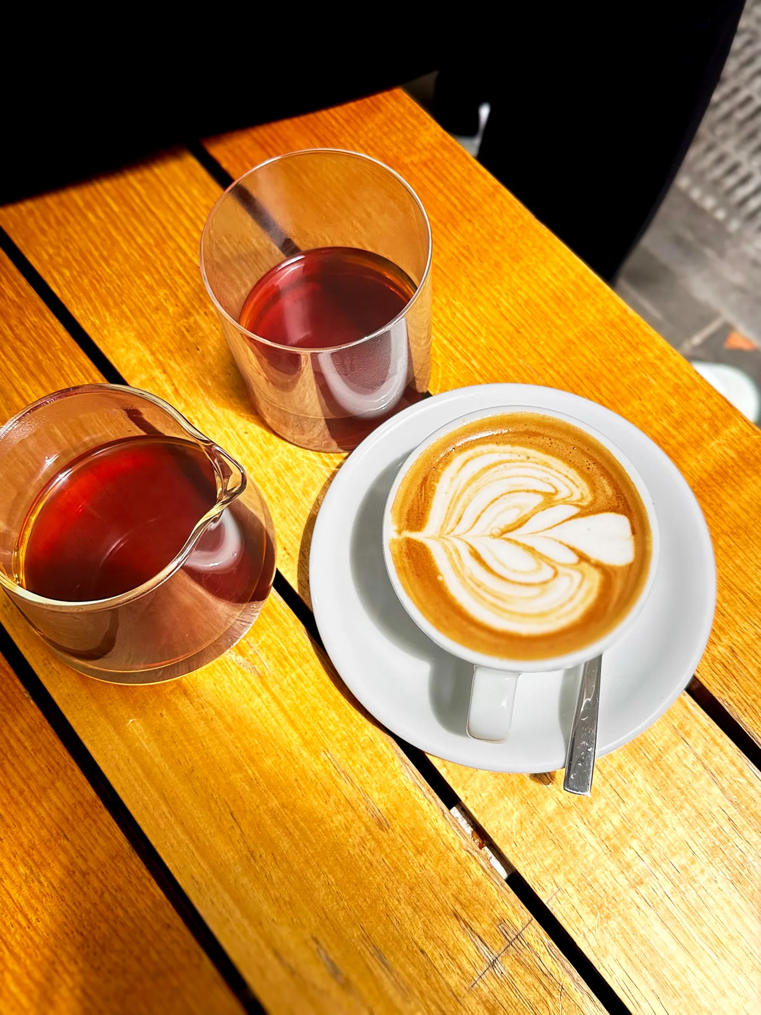 A flat white with a rosetta beside a glass carafe of filter coffee on a sunlit slatted table, from above