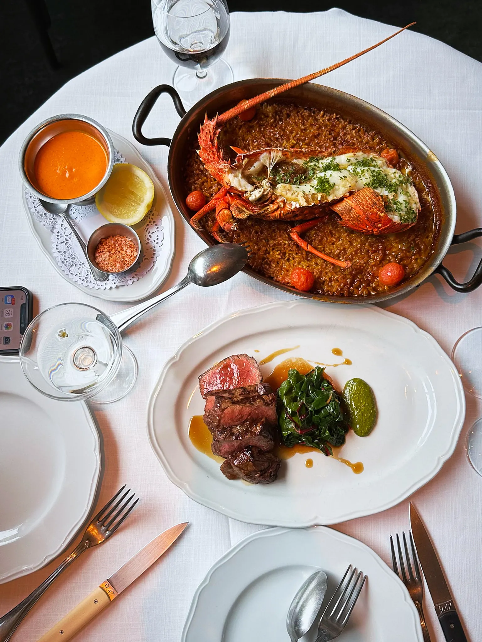 A pan of lobster rice and a plate of rare steak with greens on a white tablecloth, from above