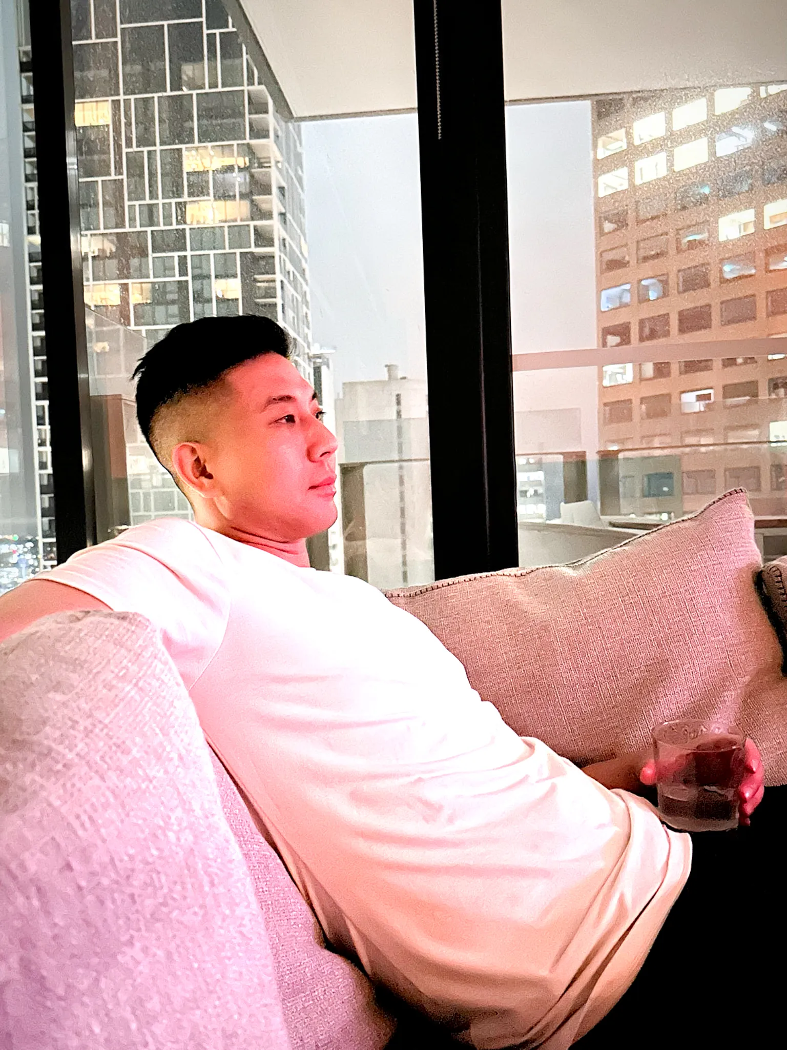 Trung on a hotel sofa by a floor-to-ceiling window, city towers behind him, a glass in his hand