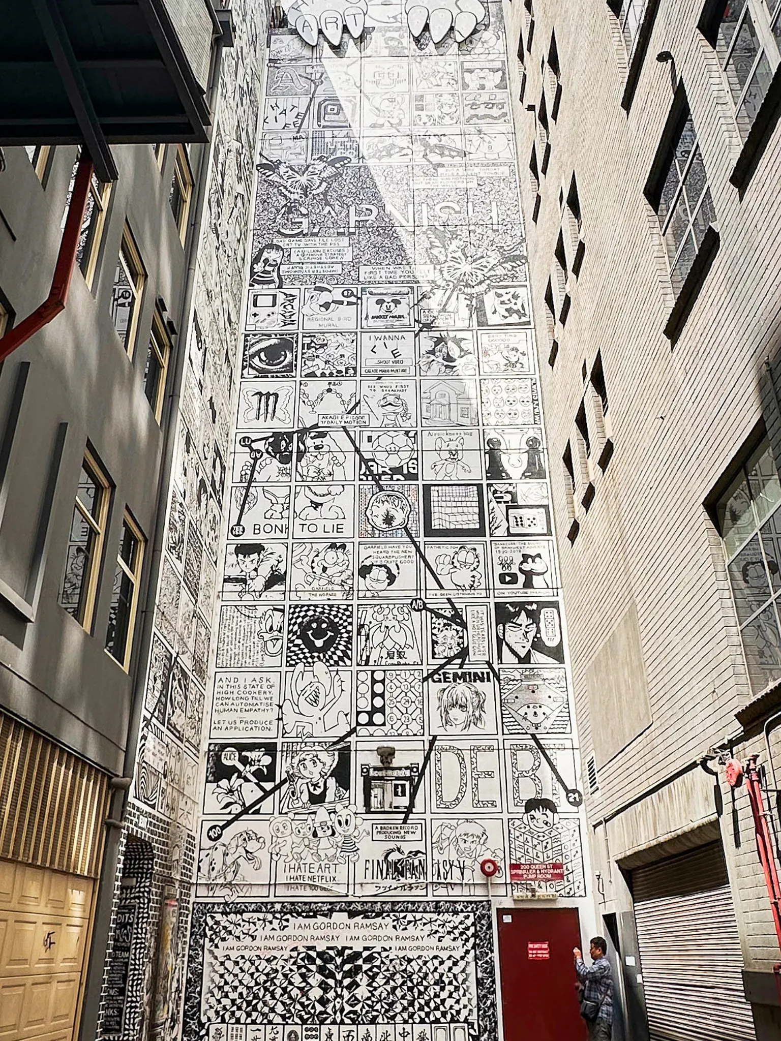 A laneway wall filled top to bottom with a black-and-white comic-panel mural, a red door at its base