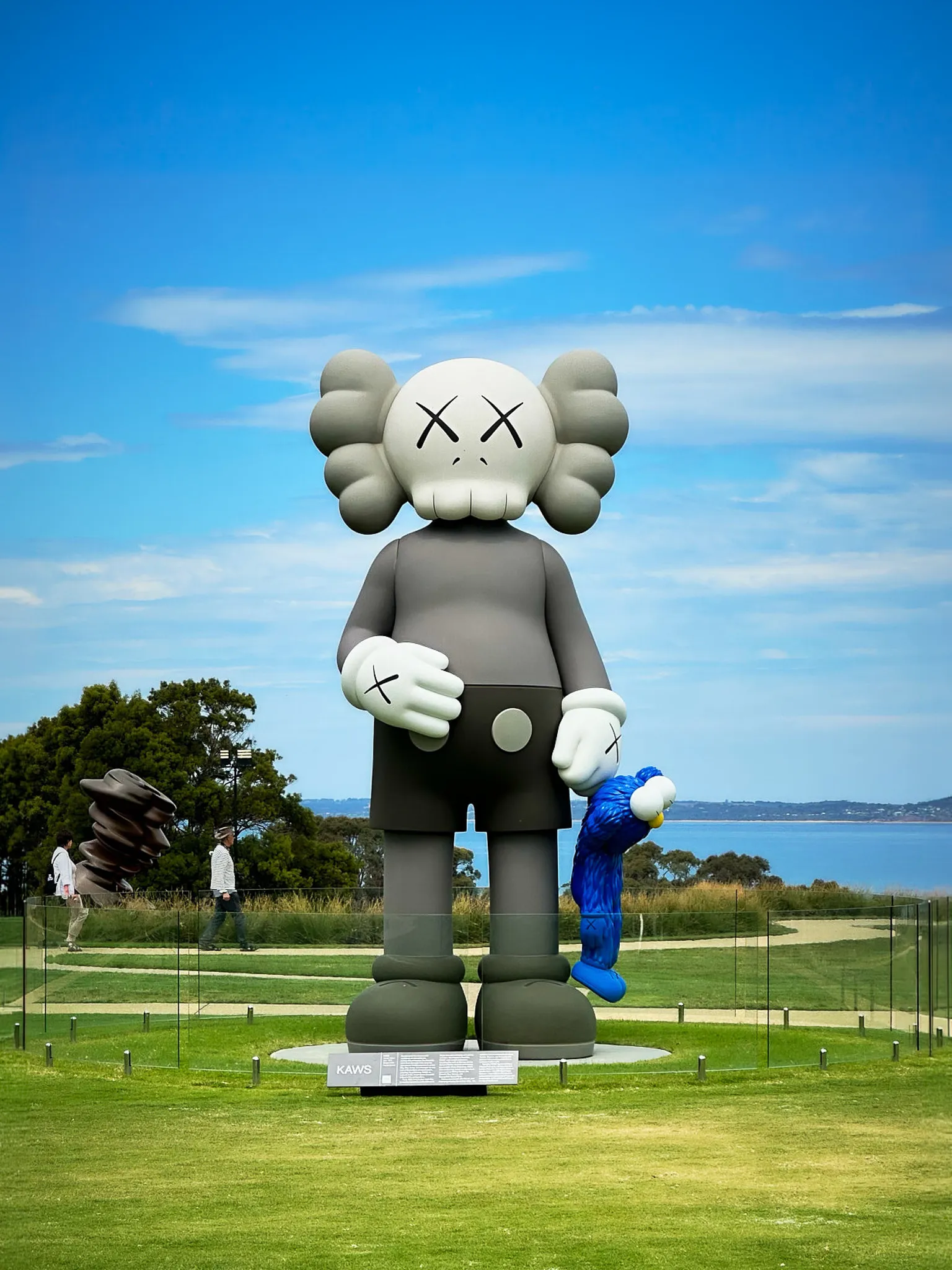 A giant grey KAWS Companion sculpture holding a small blue figure, standing on a lawn above the bay