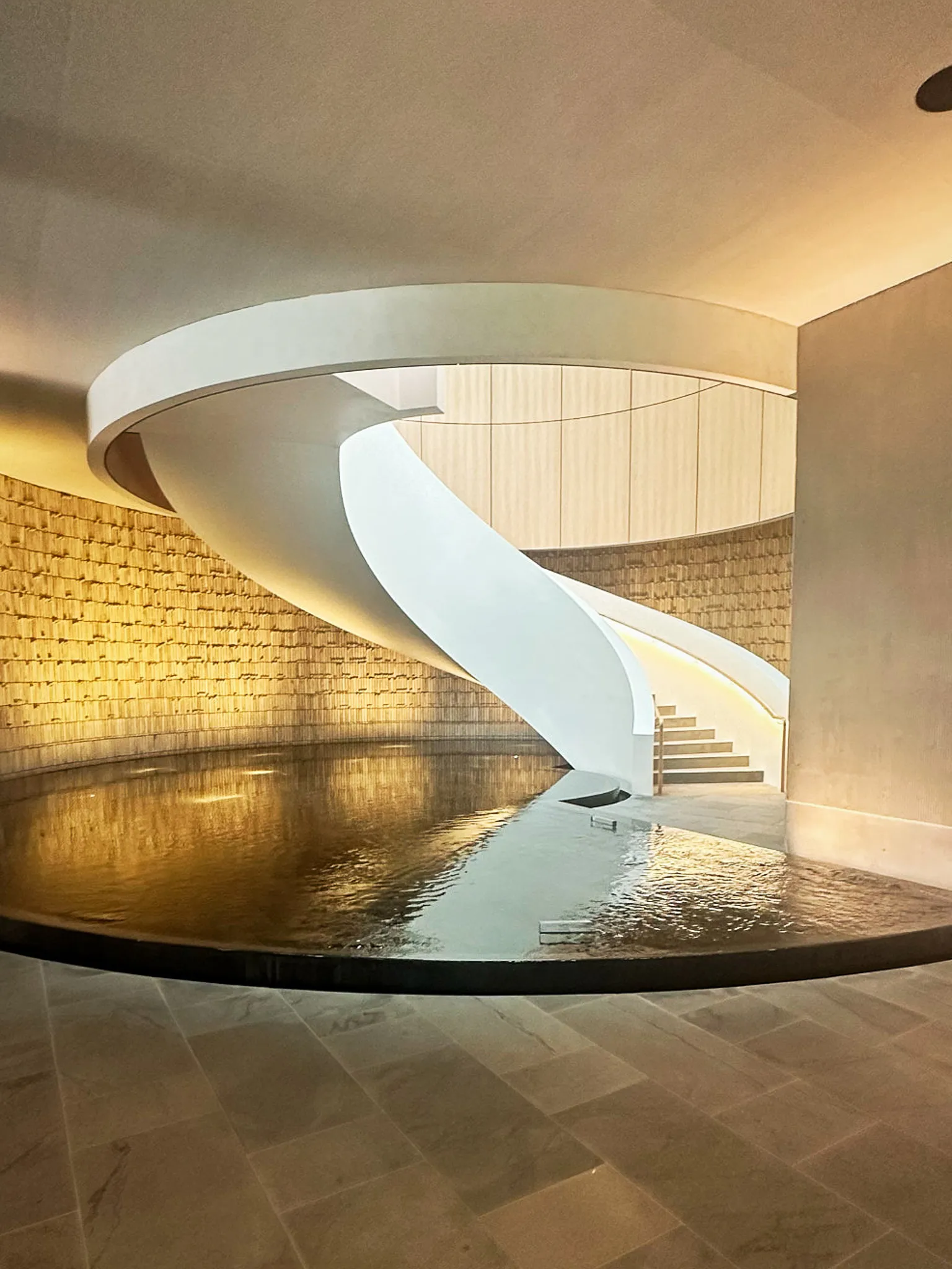 A white spiral staircase curving down to a dark reflecting pool, gold brick wall behind