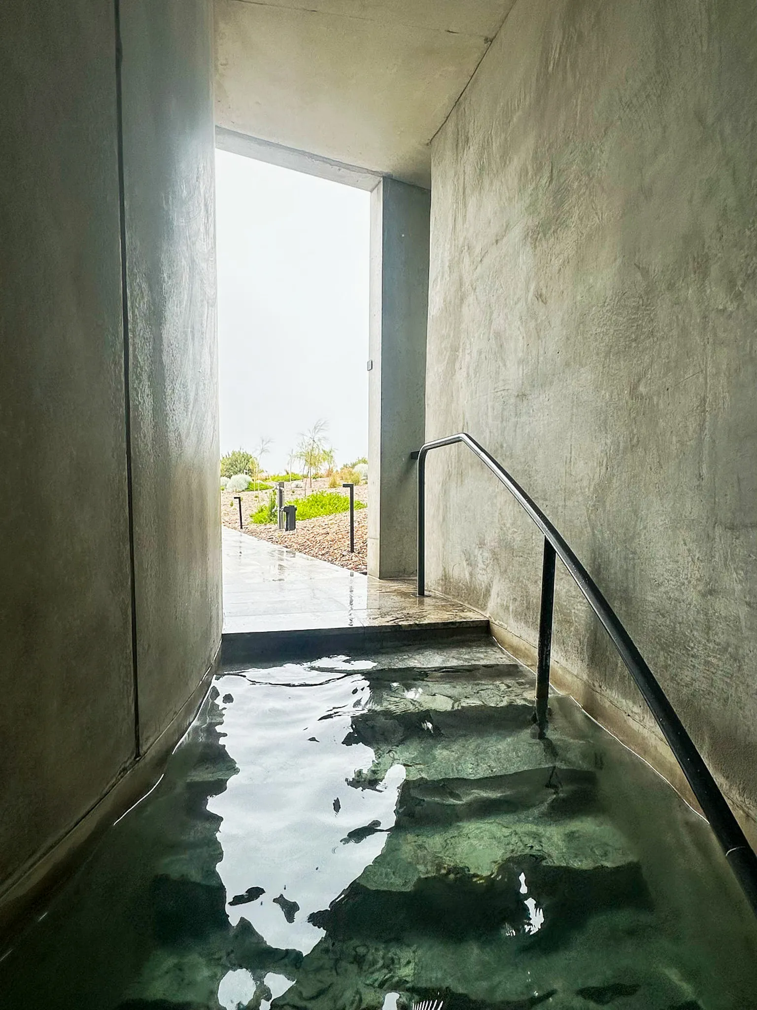 A concrete passage with steps descending into green thermal water, daylight at the far opening