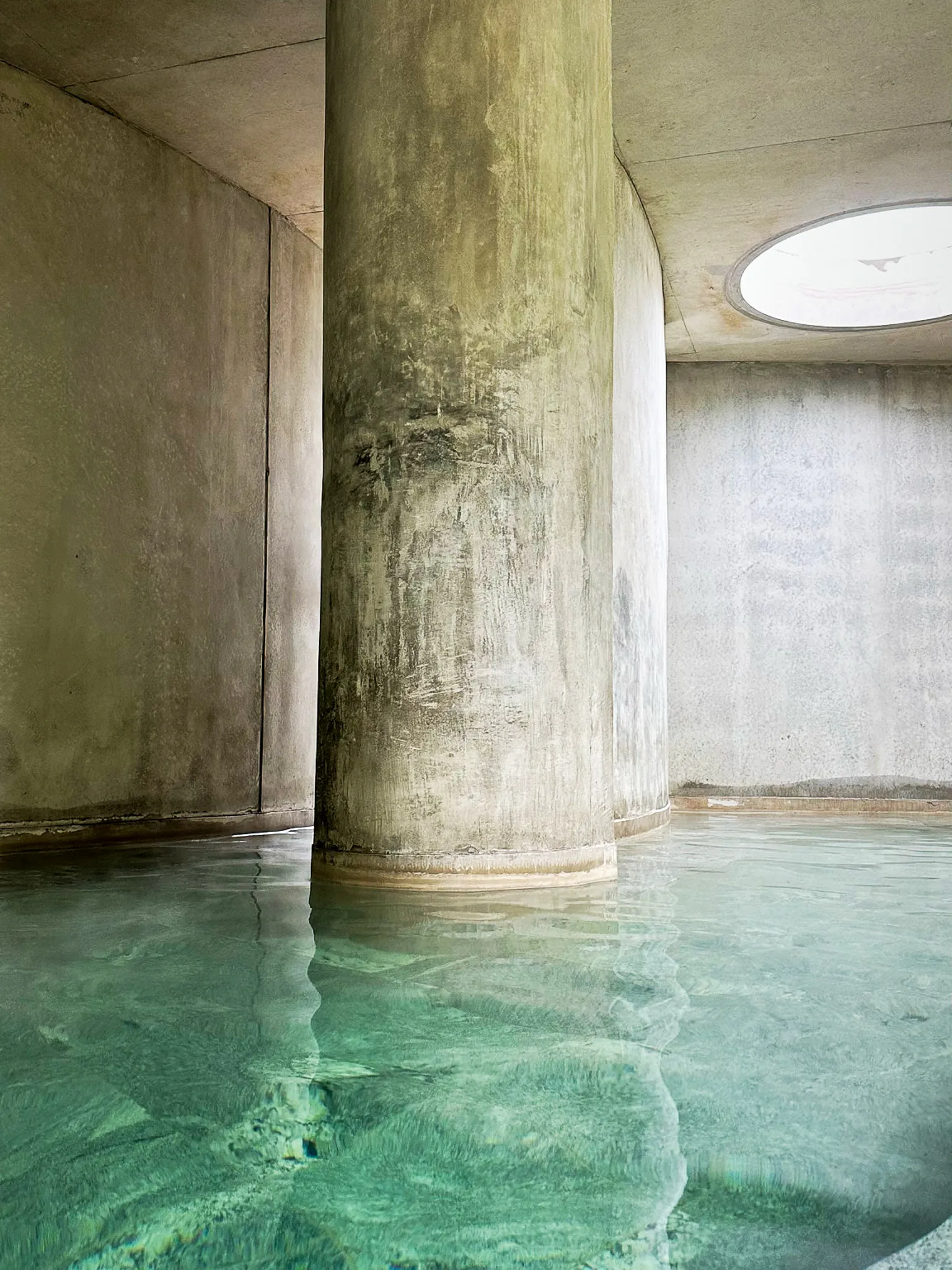 A thick concrete column standing in a pale green thermal pool, an oculus in the ceiling above