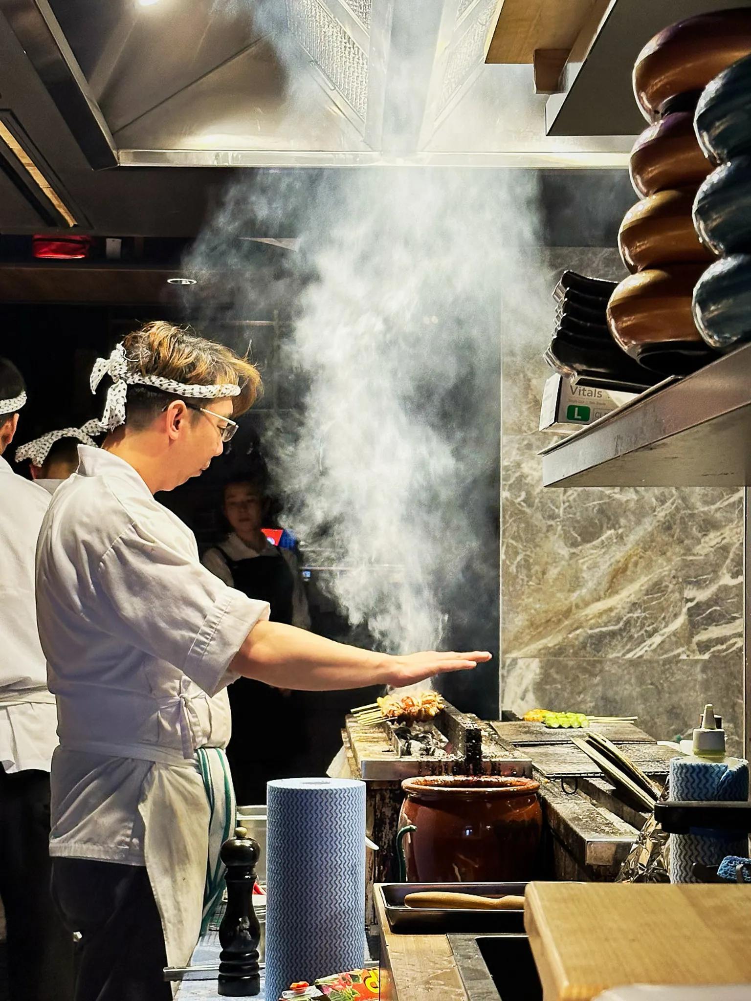 A chef in a tenugui headband working over a grill in a cloud of smoke