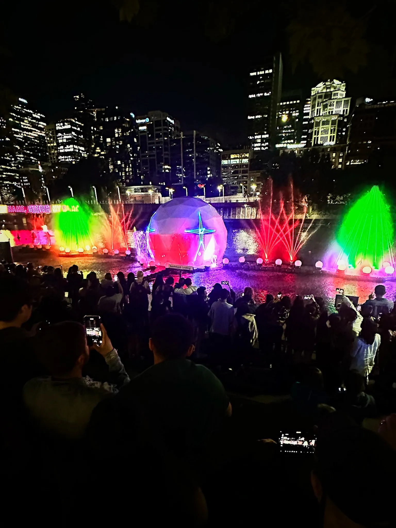 A night crowd watching a lit dome and coloured water jets over a lake, phones raised