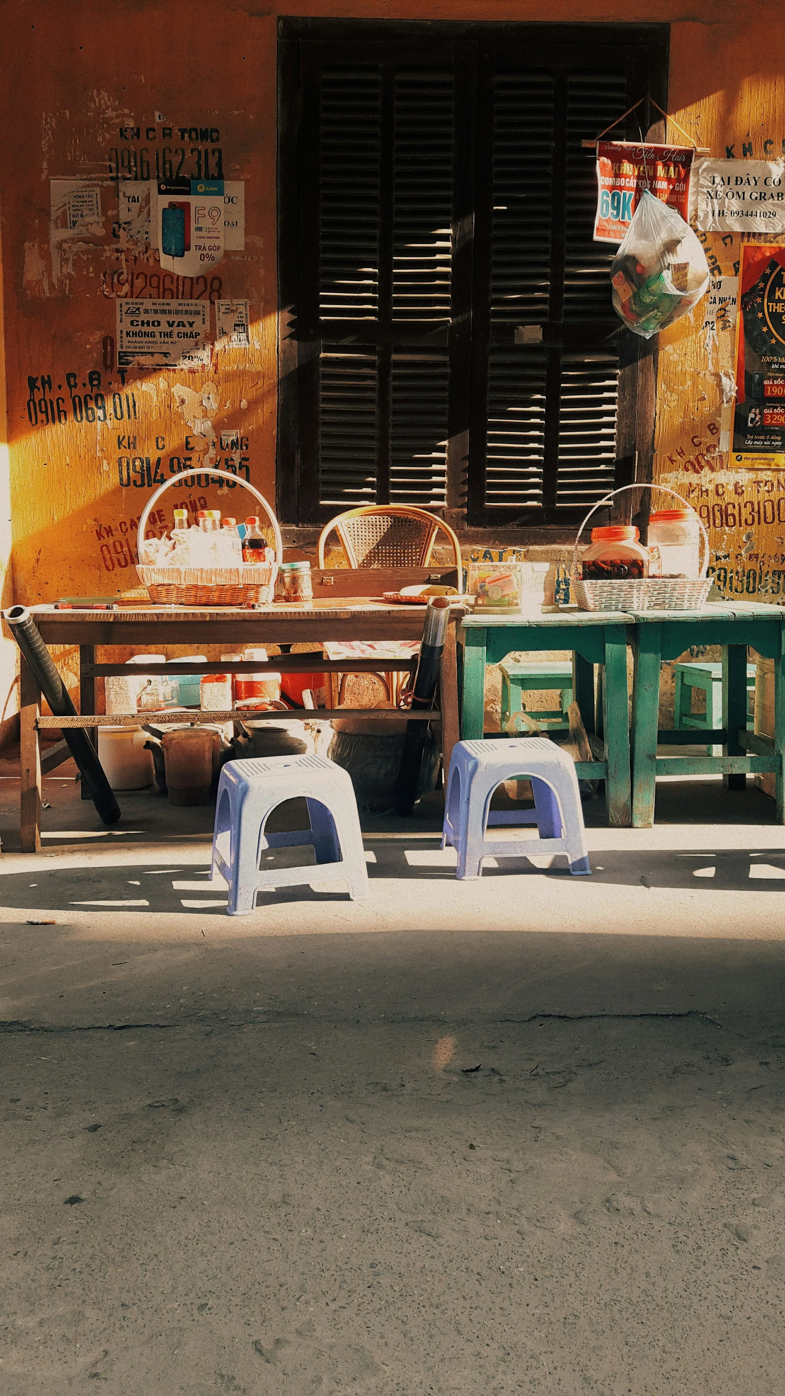 Empty low plastic stools and a table set out on the pavement in front of a mustard-yellow shopfront with dark wooden shutters